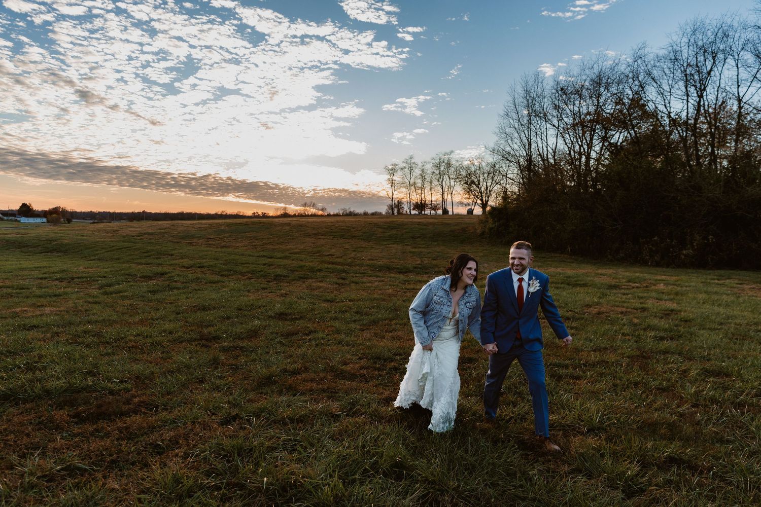 A couple shares a playful moment in an open field at sunset with trees silhouetted in the background.
