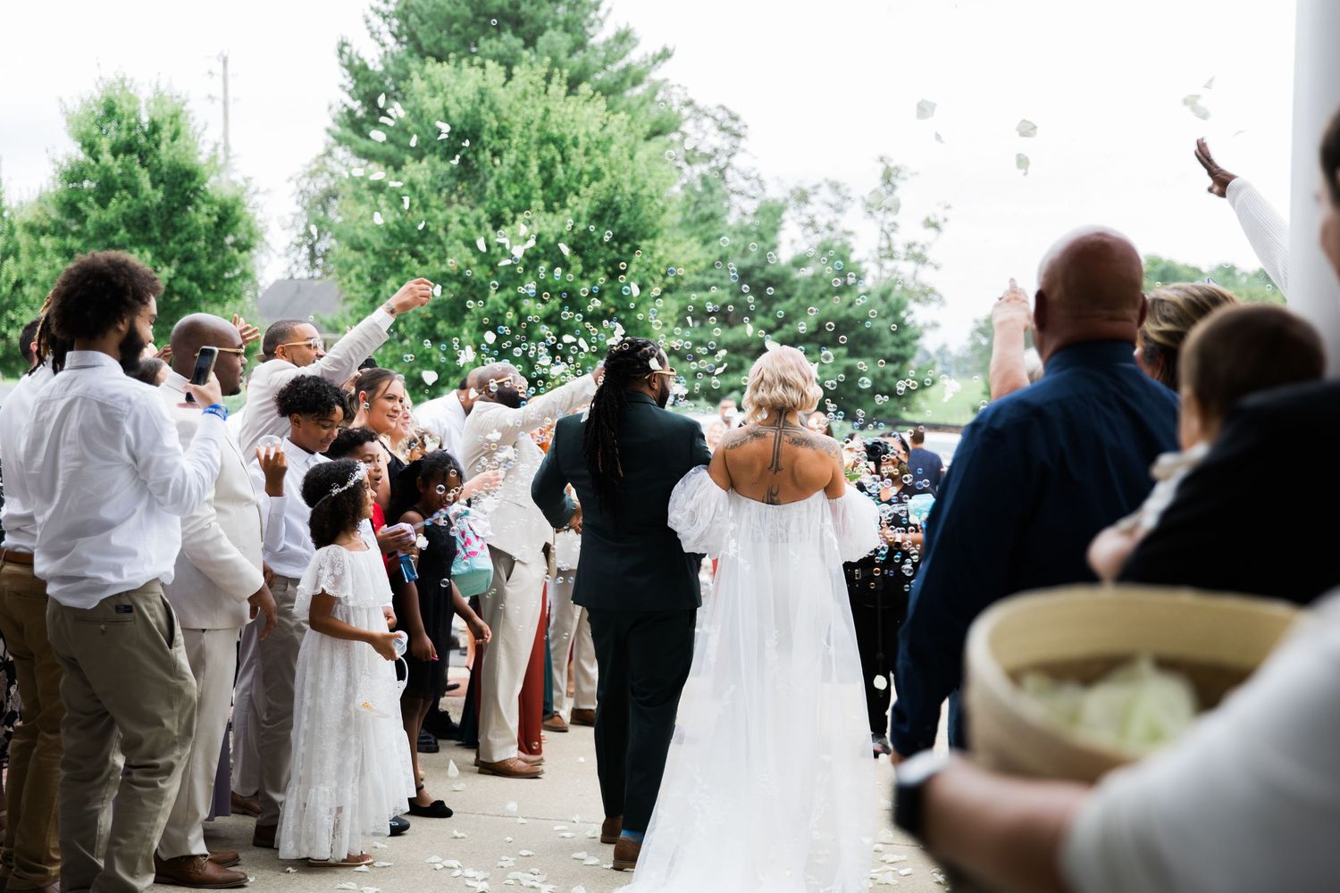 Wedding guests line a path to celebrate as a couple exits amid flying paper confetti on a sunny afternoon.