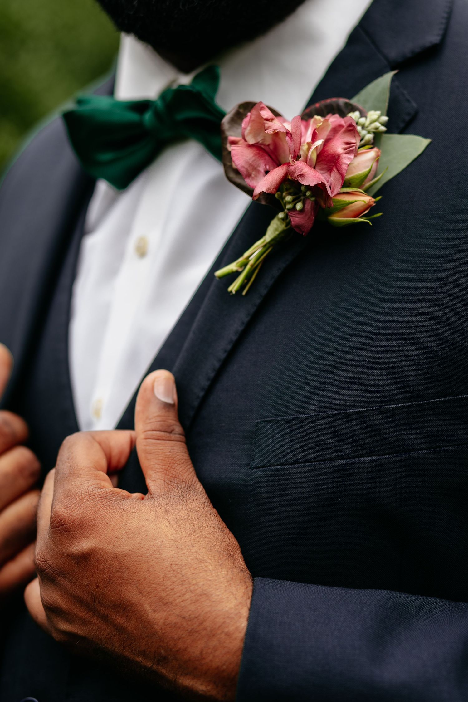 Close-up of groom's boutonniere with pink carnation and emerald green bow tie on navy suit lapel.