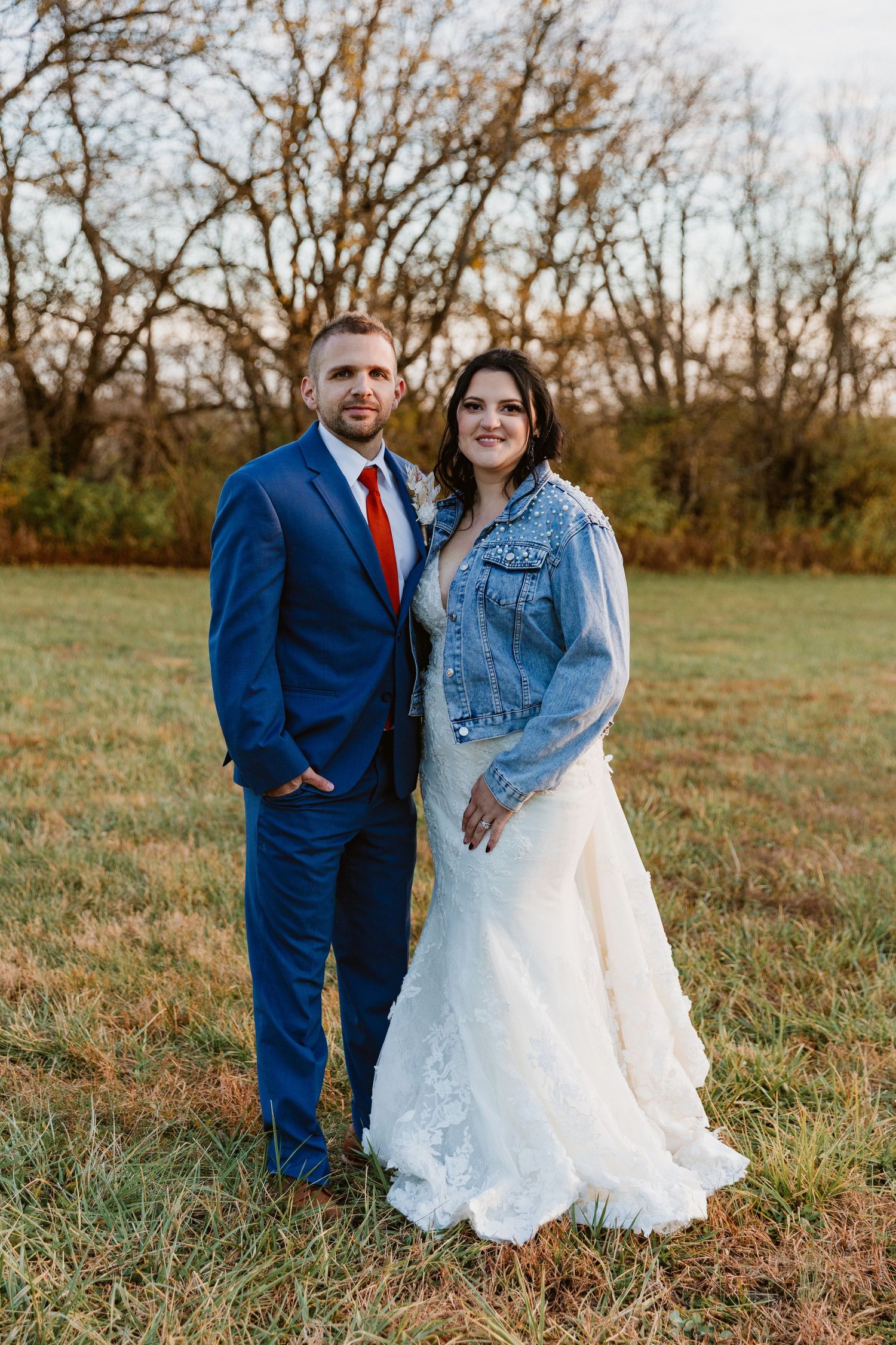 A couple poses outdoors in wedding attire, with the bride wearing a denim jacket over her lace gown and groom in navy suit.