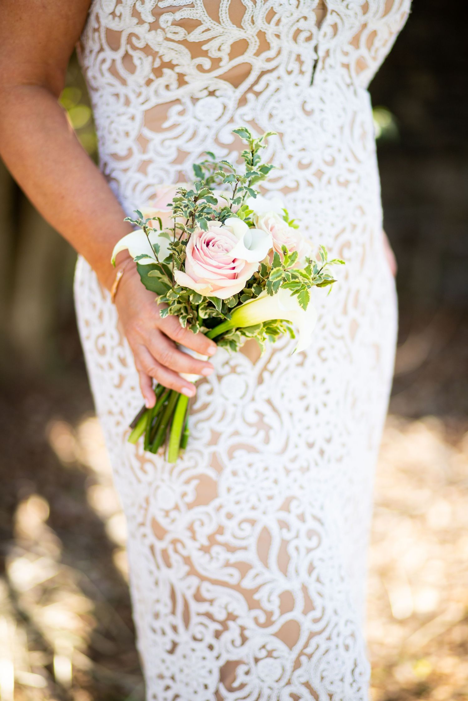 A delicate pink rose bouquet held against an intricate white lace wedding dress in sunlit outdoor setting.