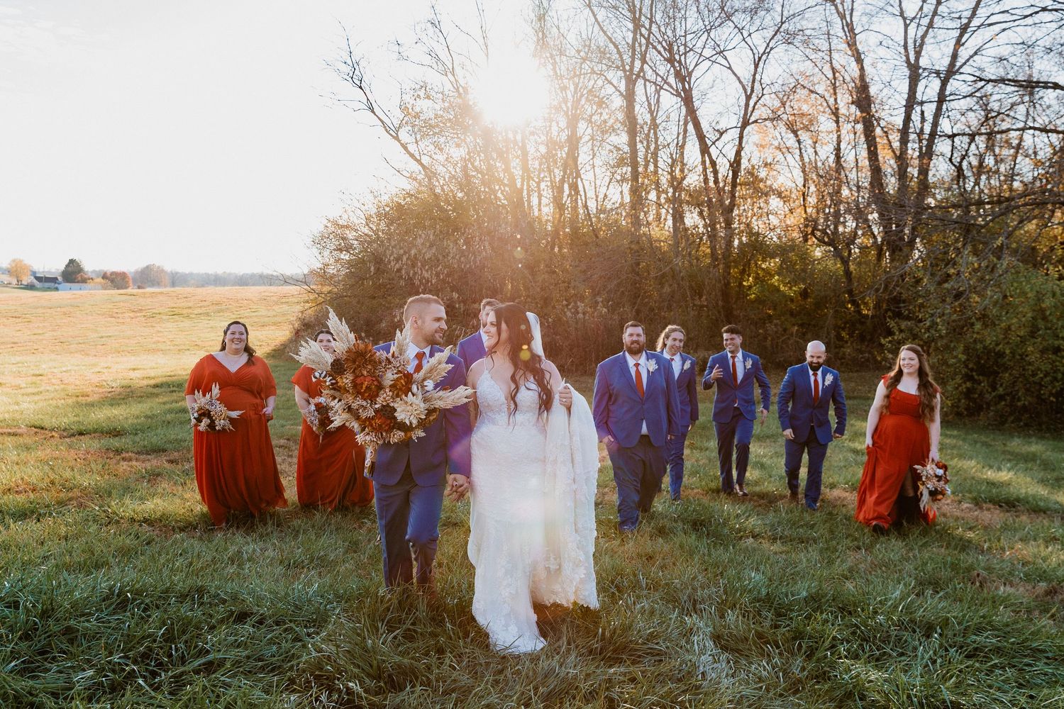 A wedding party poses together in a sunlit field during golden hour with bridesmaids in red and groomsmen in purple.