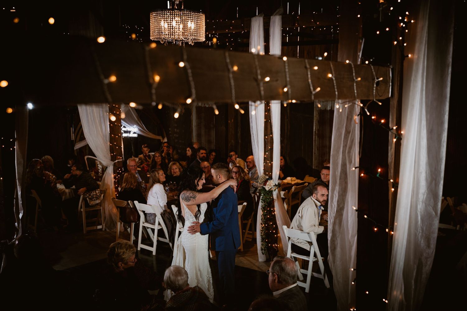 Wedding reception venue decorated with string lights and white drapes as guests celebrate on the dance floor.