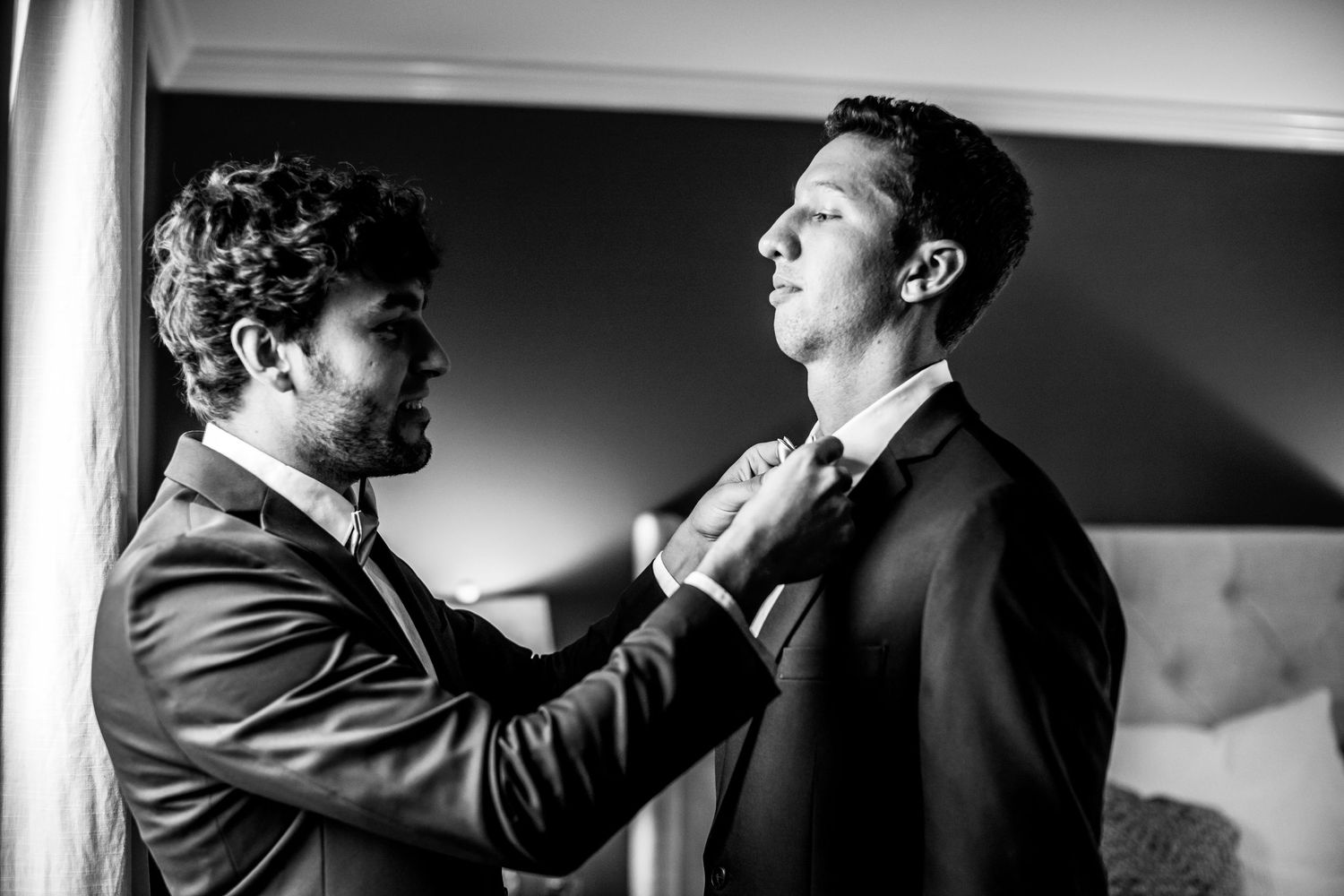Black and white photograph shows someone adjusting another person's tie in an intimate pre-wedding moment.