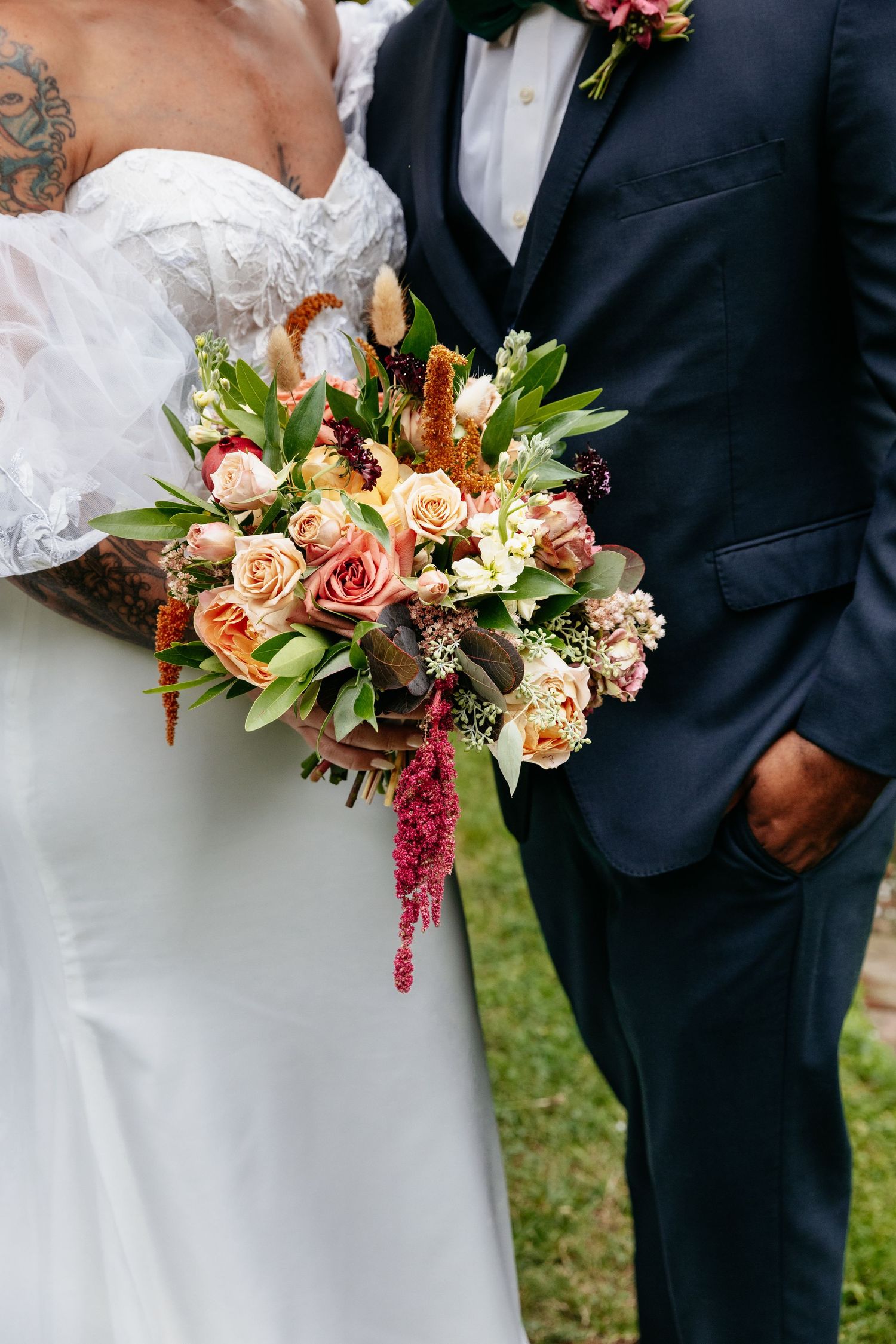 Close up wedding photo showing an elegant bouquet with peach roses and hanging amaranth between a white dress and navy suit.