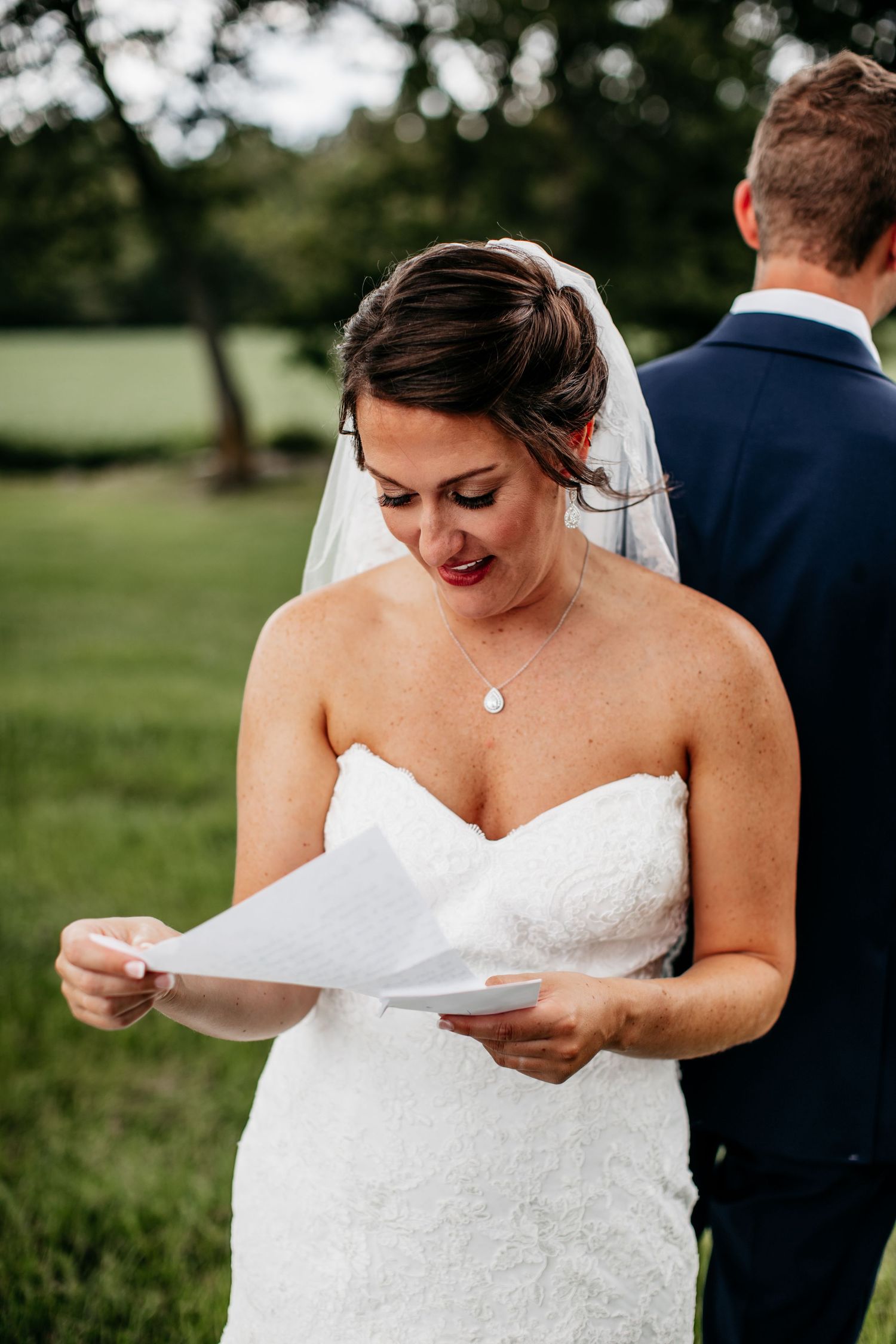 Bride in a strapless lace wedding dress reads a letter outdoors while standing in a grassy field.