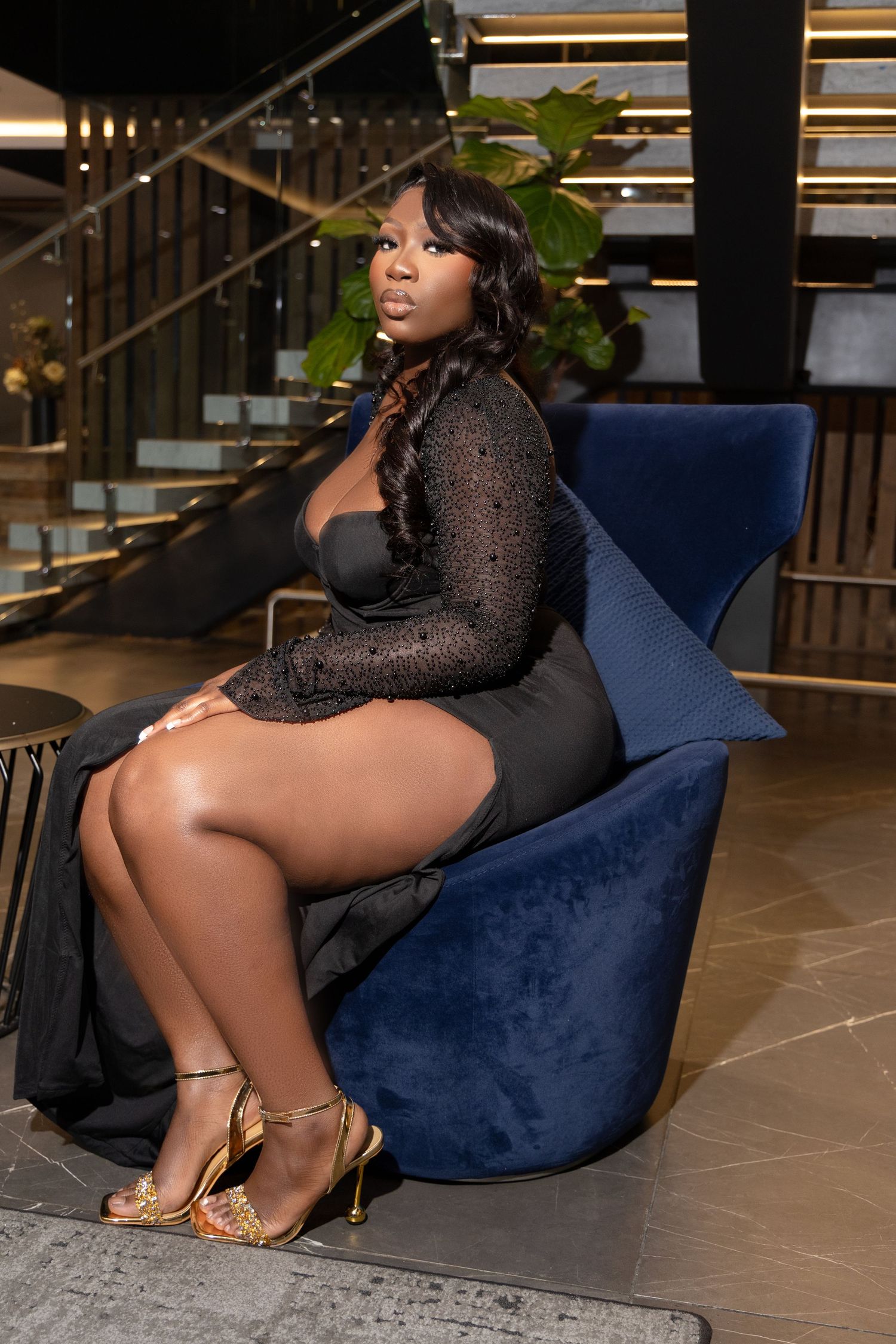 Elegant black evening gown and gold heels styled with modern blue velvet chair in upscale setting.