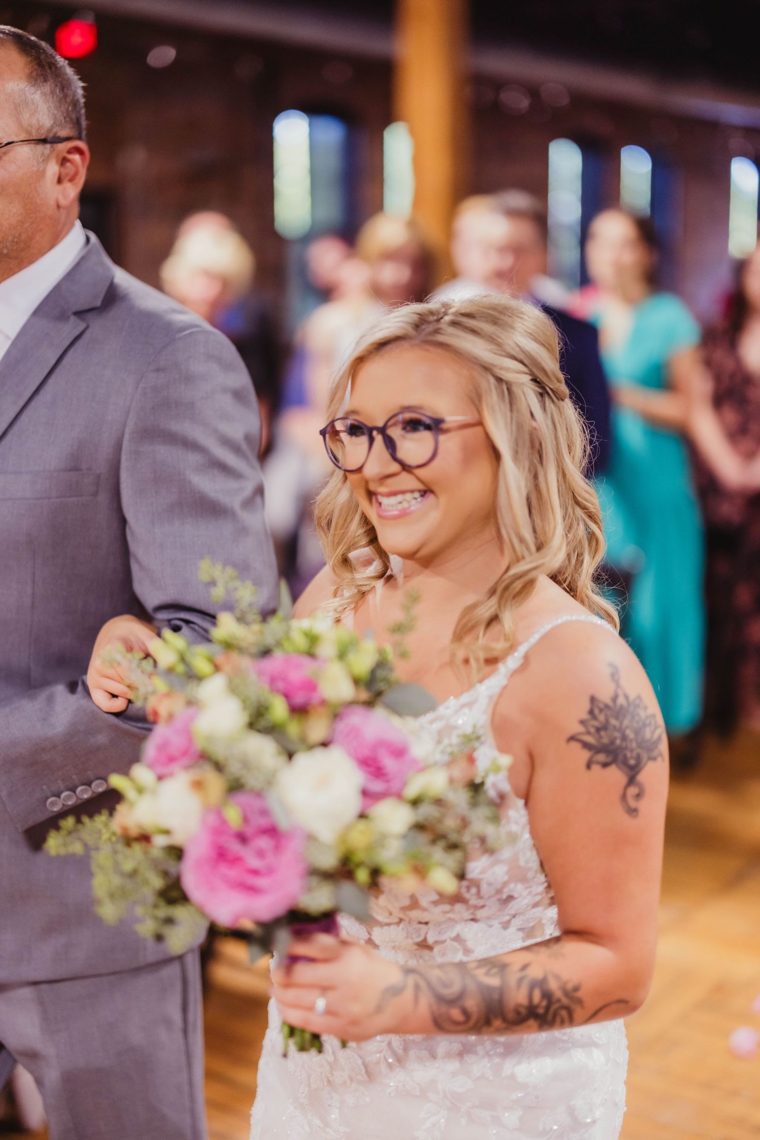Bride holding pink and white bouquet smiles during wedding ceremony in rustic venue with exposed brick walls.