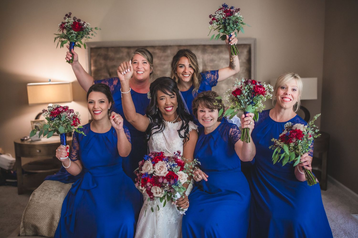 Joyful bridesmaids in blue lift bouquets alongside bride in white gown.