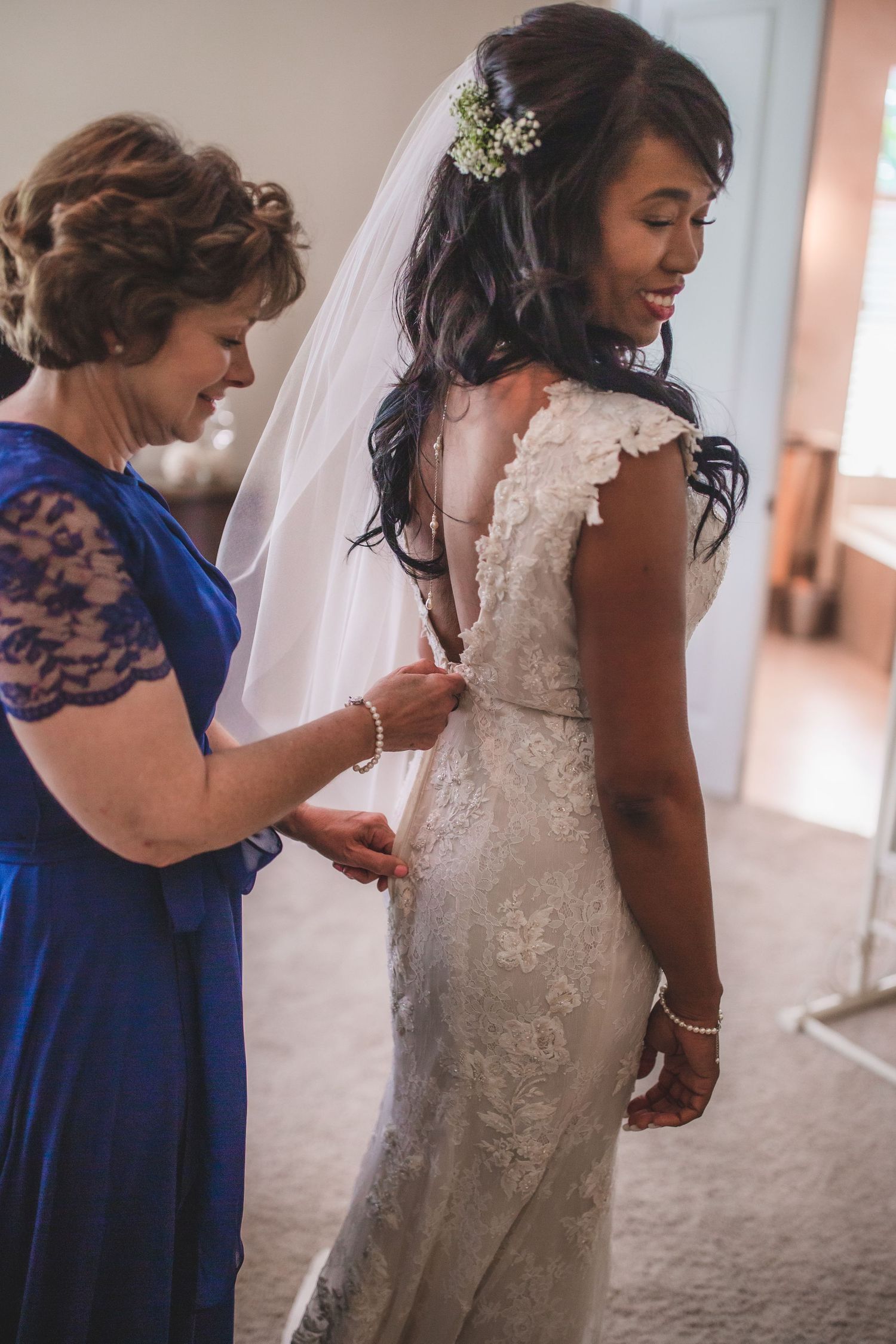 Final adjustments being made to a bride's fitted lace wedding dress by a helper in royal blue.