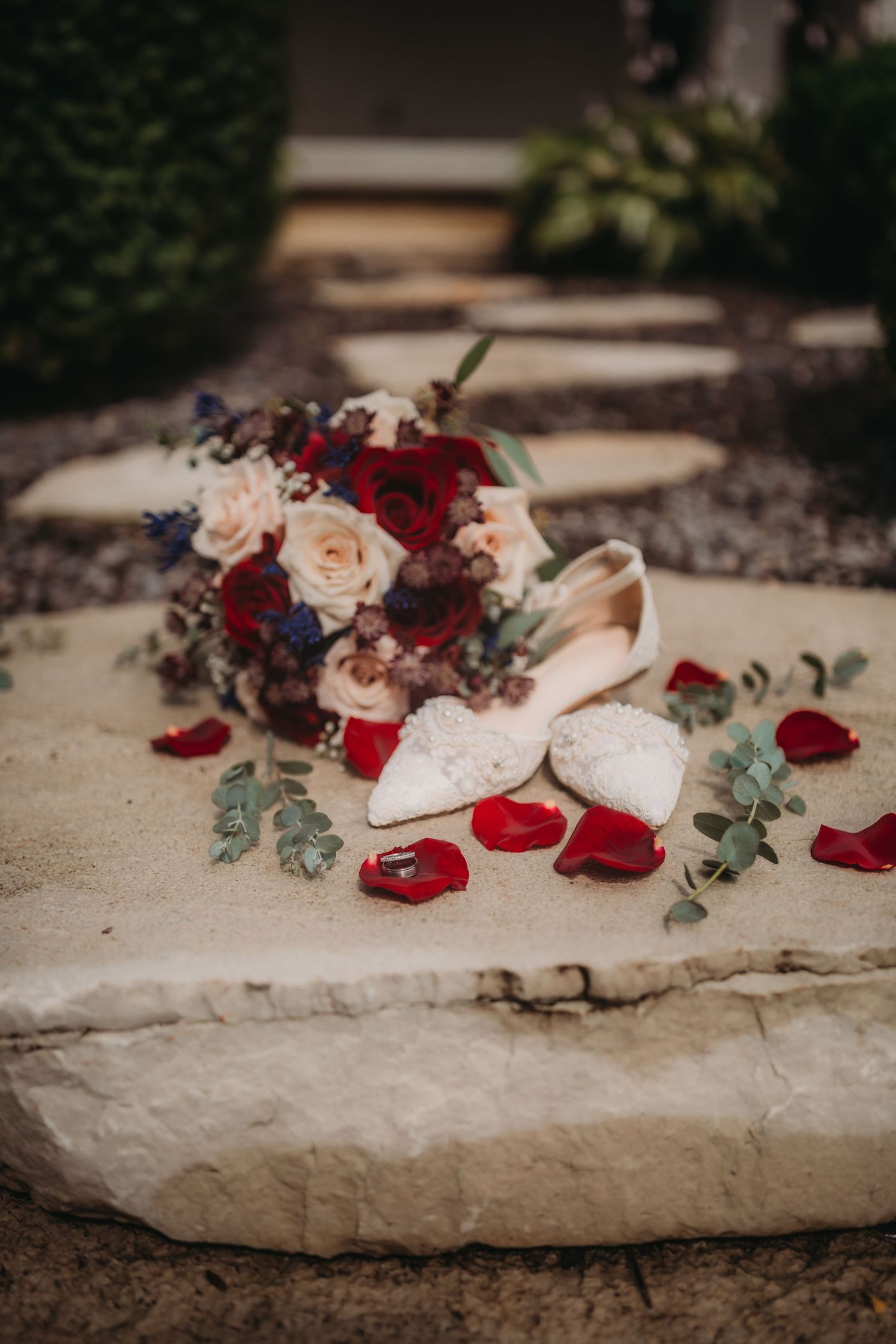 Wedding bouquet with red and blush roses next to white lace shoes on stone steps.