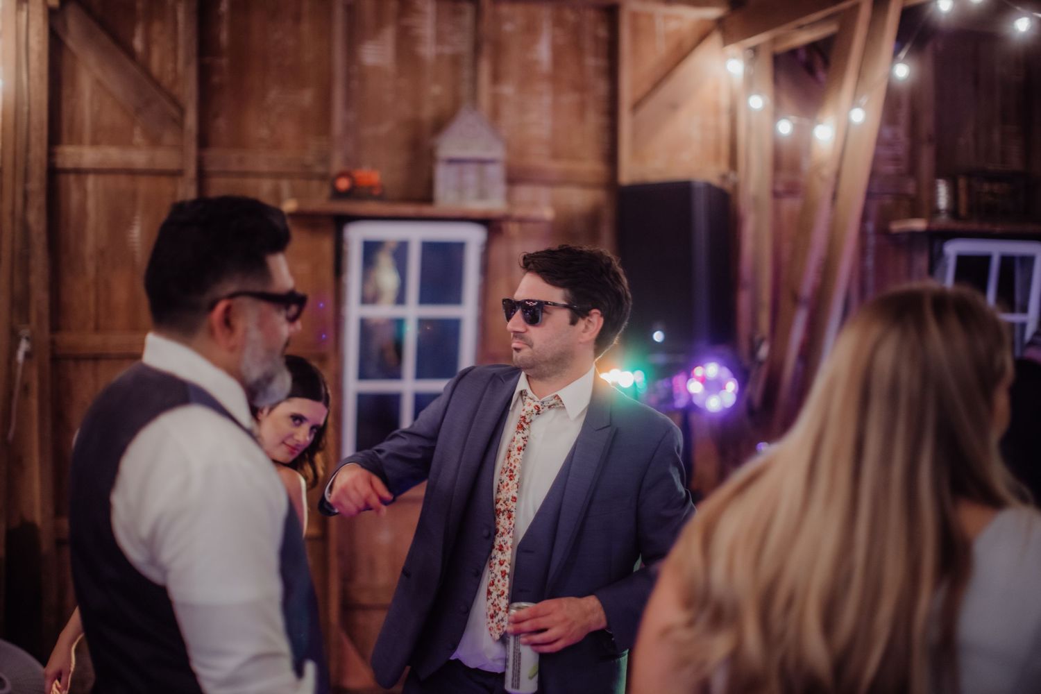 Wedding guests dance and celebrate in a warmly lit rustic barn venue with string lights overhead.