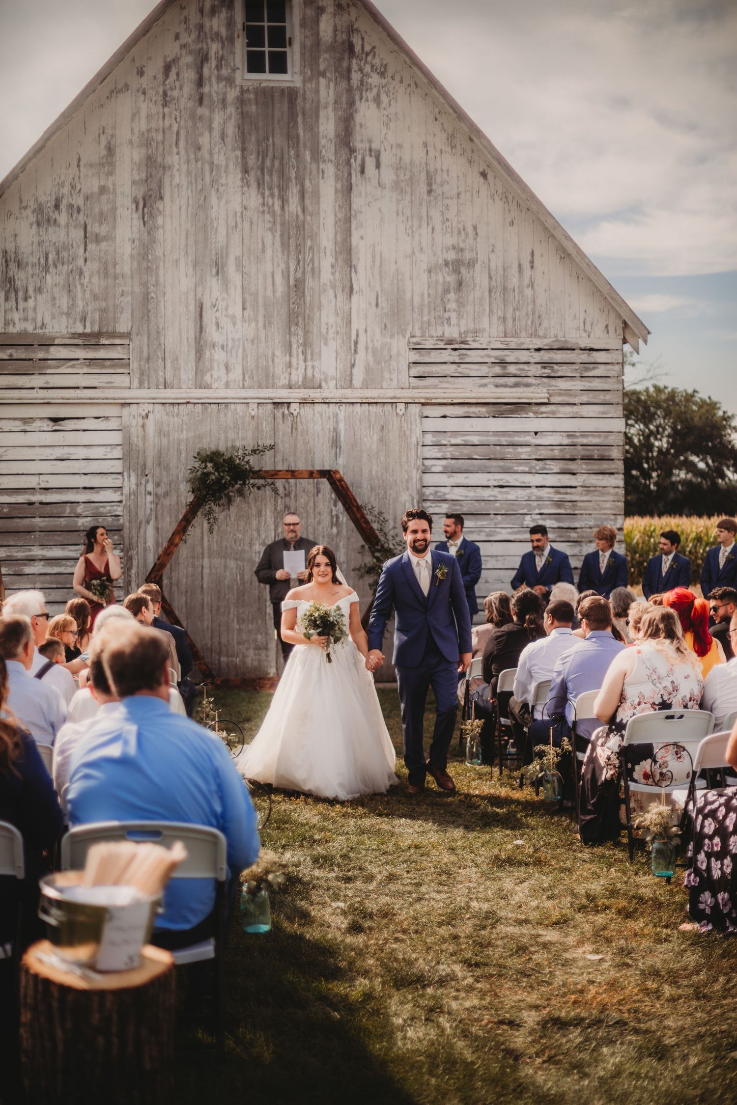 Wedding ceremony in progress outside vintage white barn with guests seated on folding chairs.