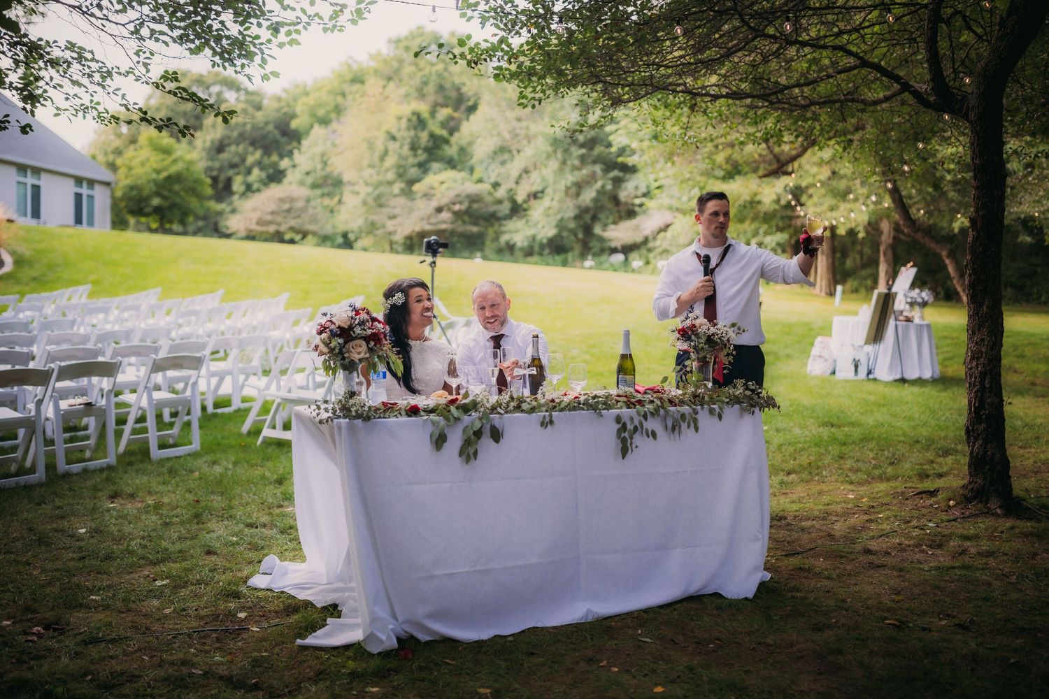 Outdoor wedding reception setup with long white tables decorated with floral arrangements under shady trees.