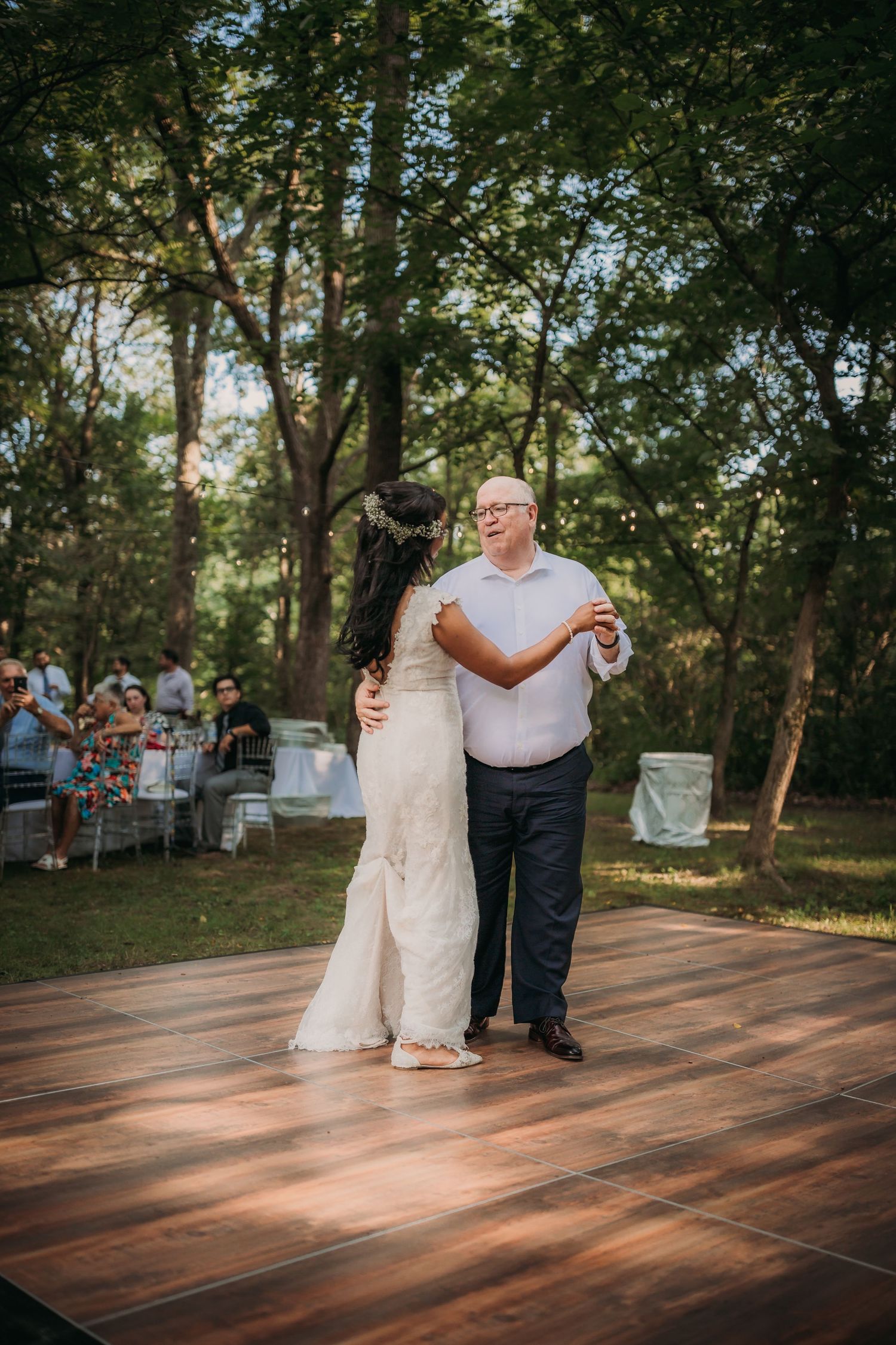 Wedding couple dances together on a wooden platform under trees with string lights and guests watching.