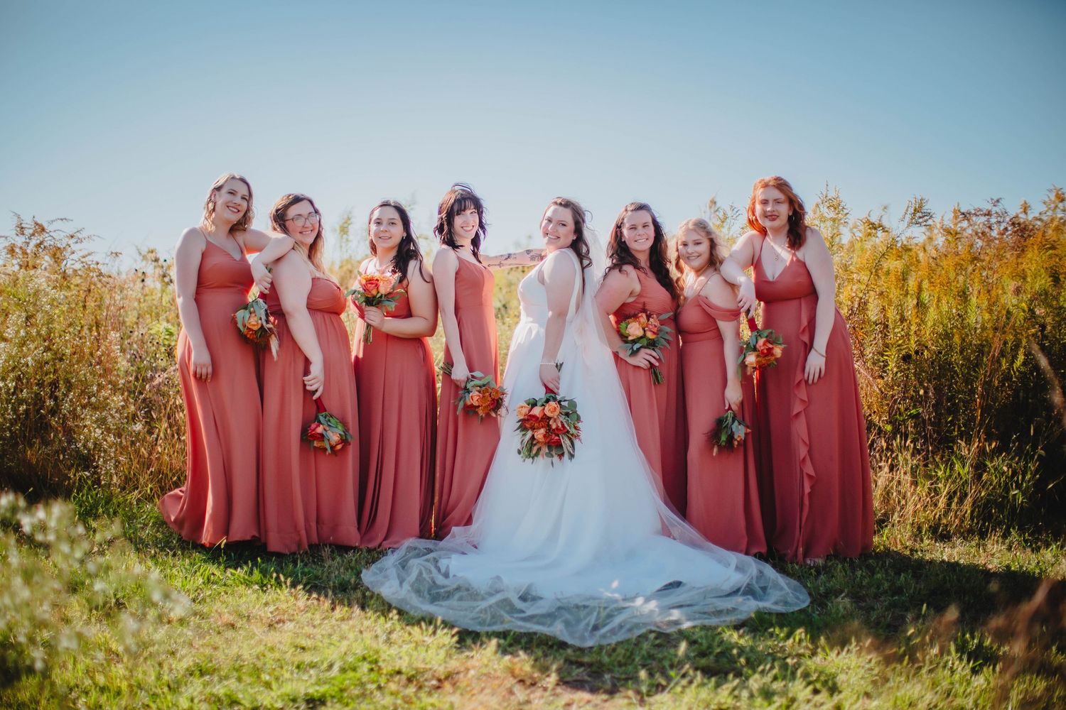 Bridal party wearing coral dresses poses with bride in white gown against golden autumn field.