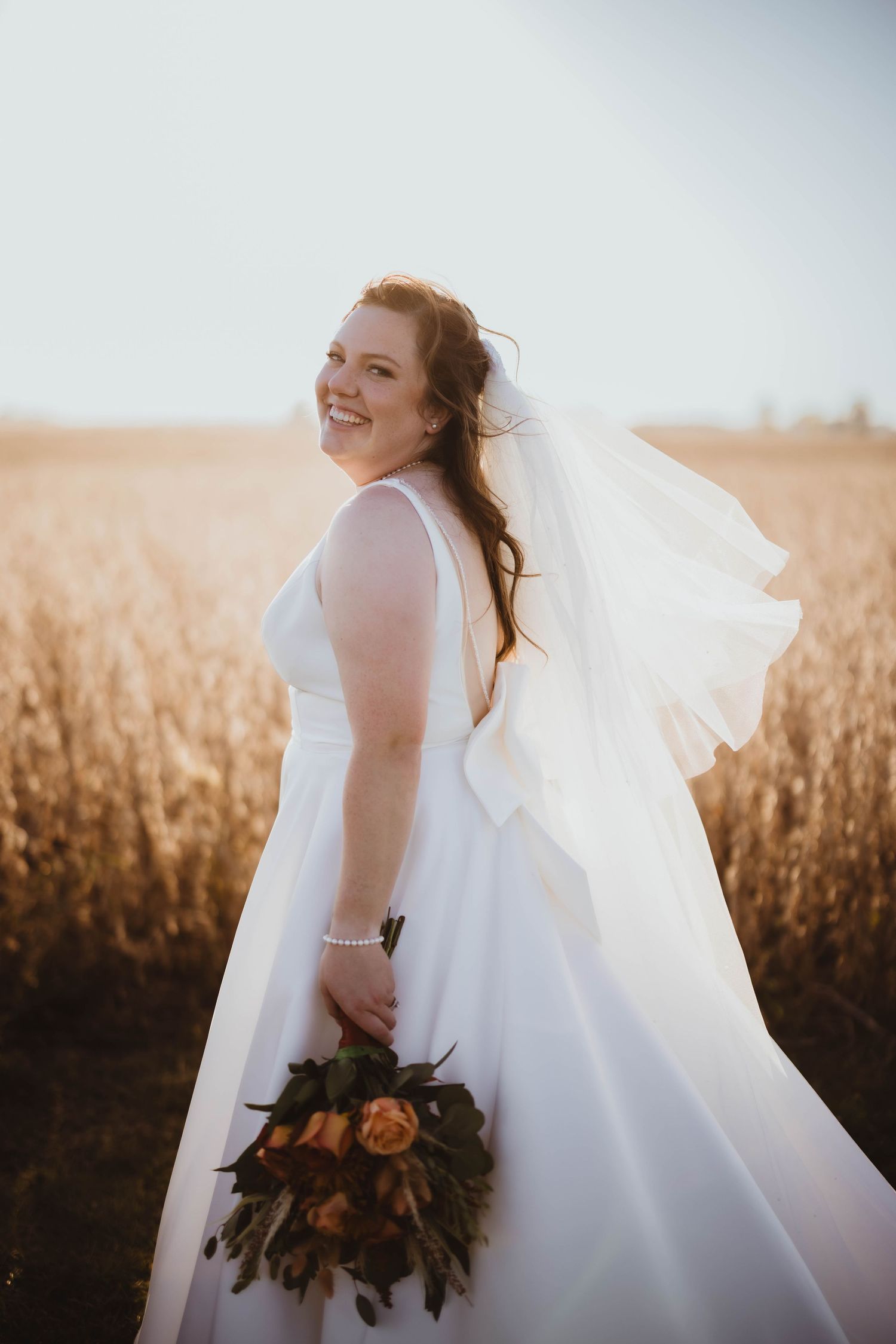 Bride in elegant white dress with autumn bouquet posing in golden field.
