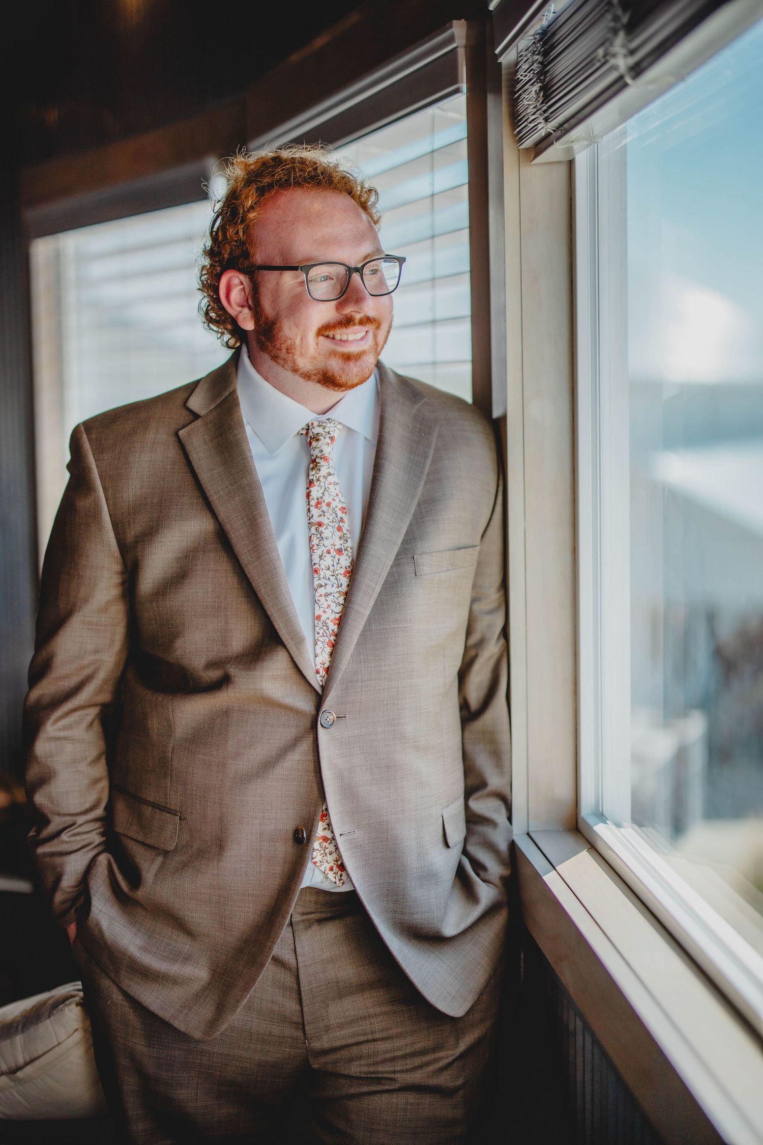 Someone in a tailored suit smiles while posing next to a sunlit window.
