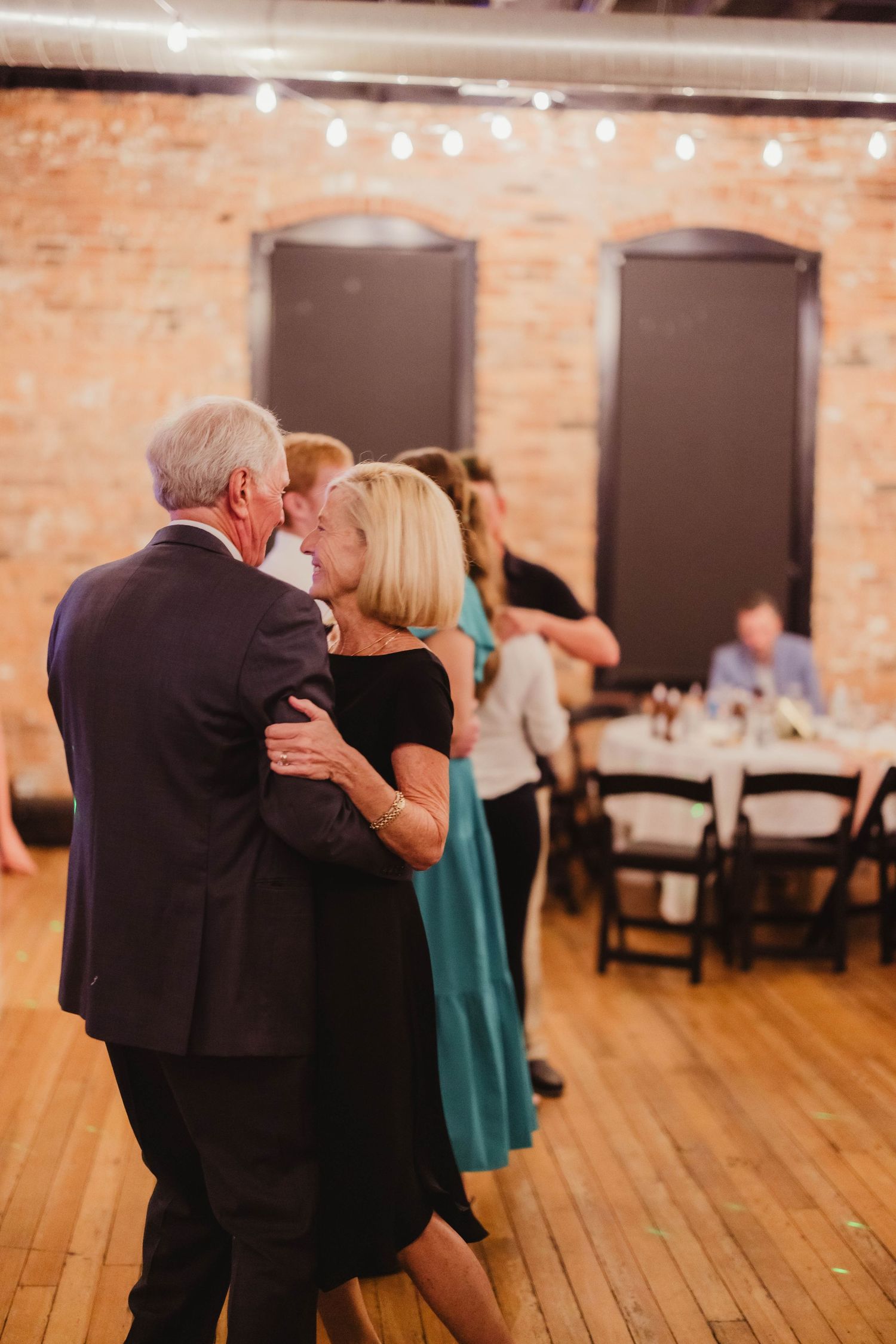 A couple dances together on a wooden floor at an event with exposed brick walls and string lights overhead.