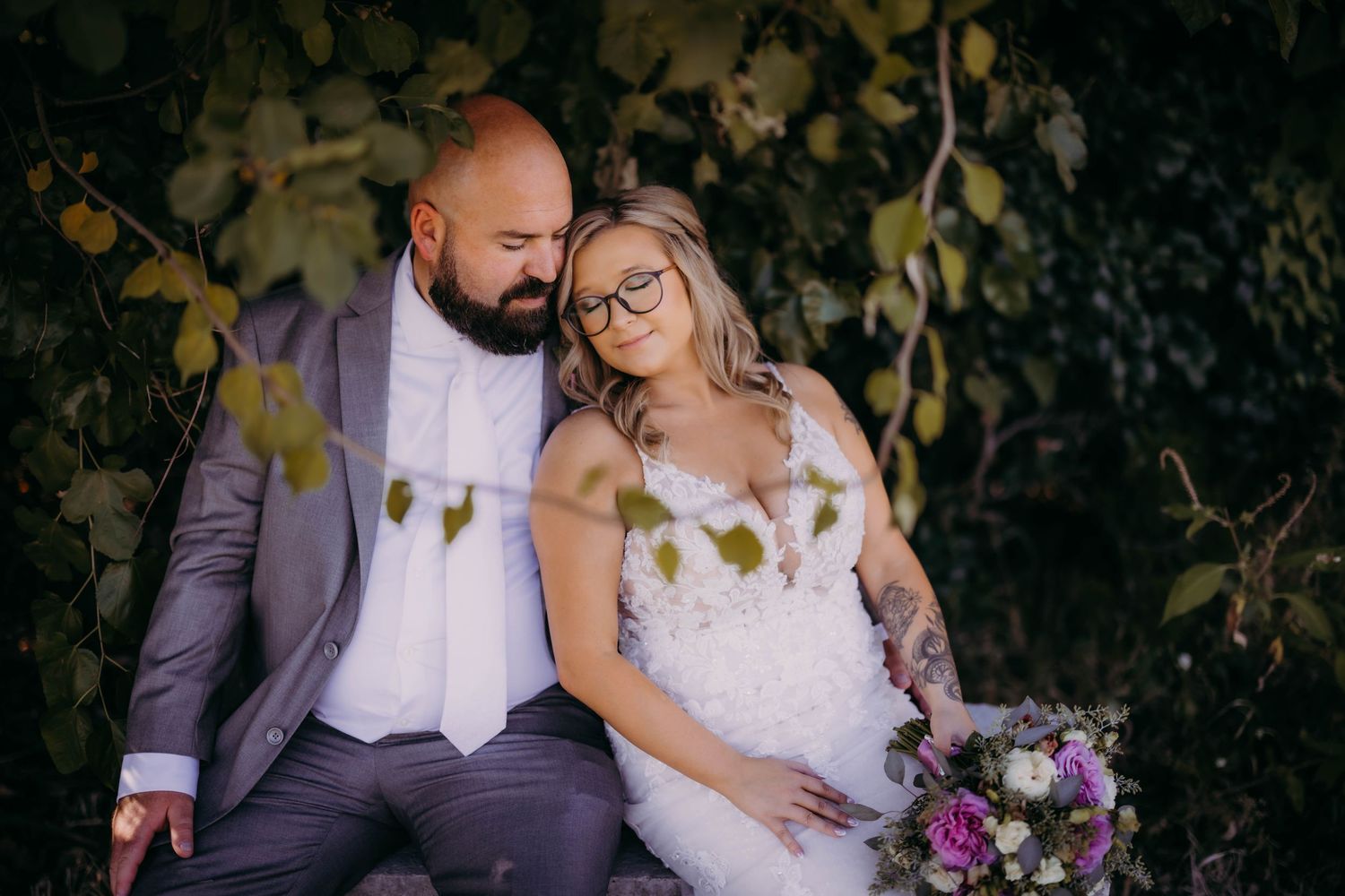 Intimate wedding portrait against lush greenery backdrop with purple floral bouquet.