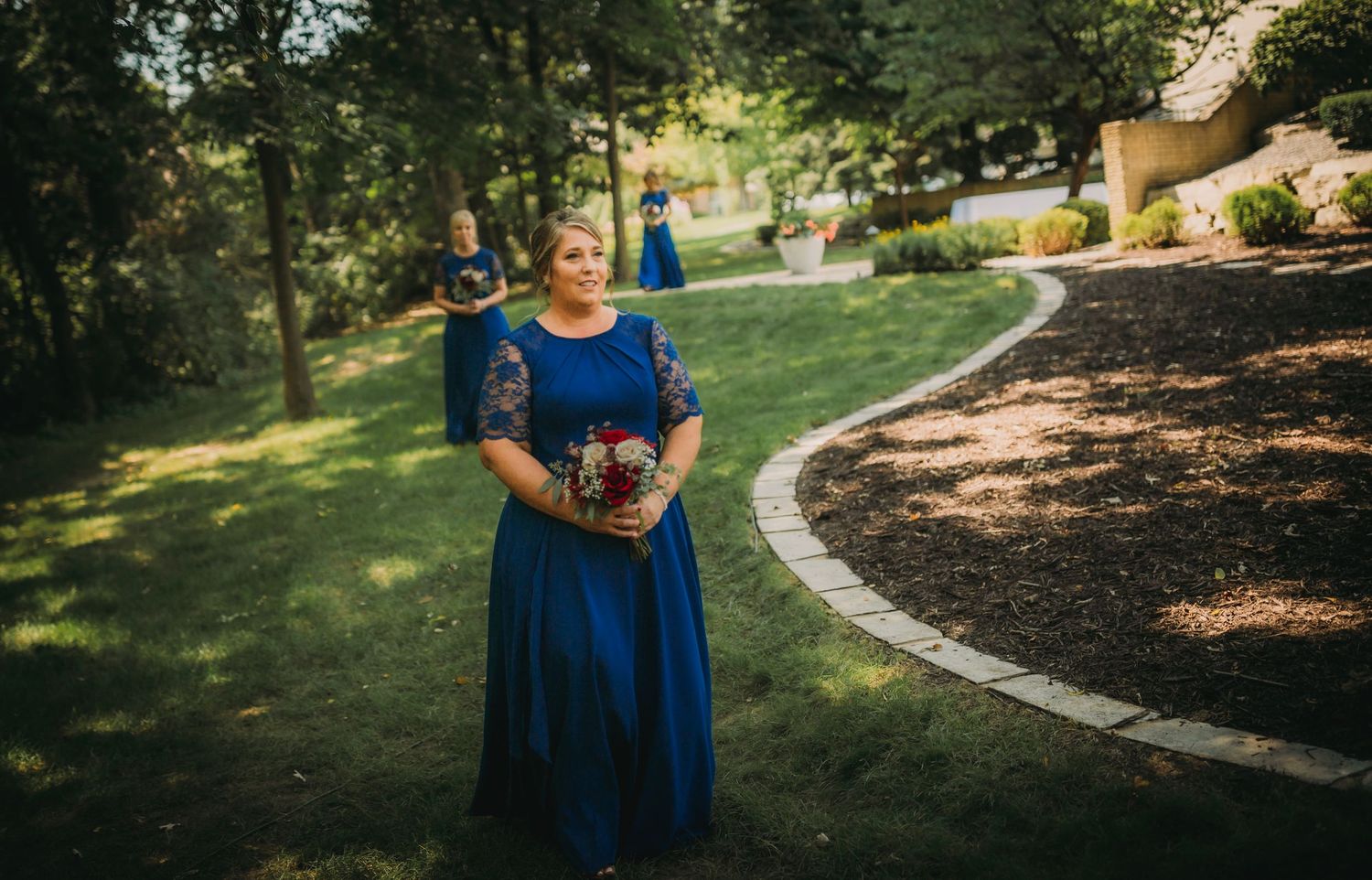 Bridesmaid in blue dress holds bouquet while standing on landscaped garden path.