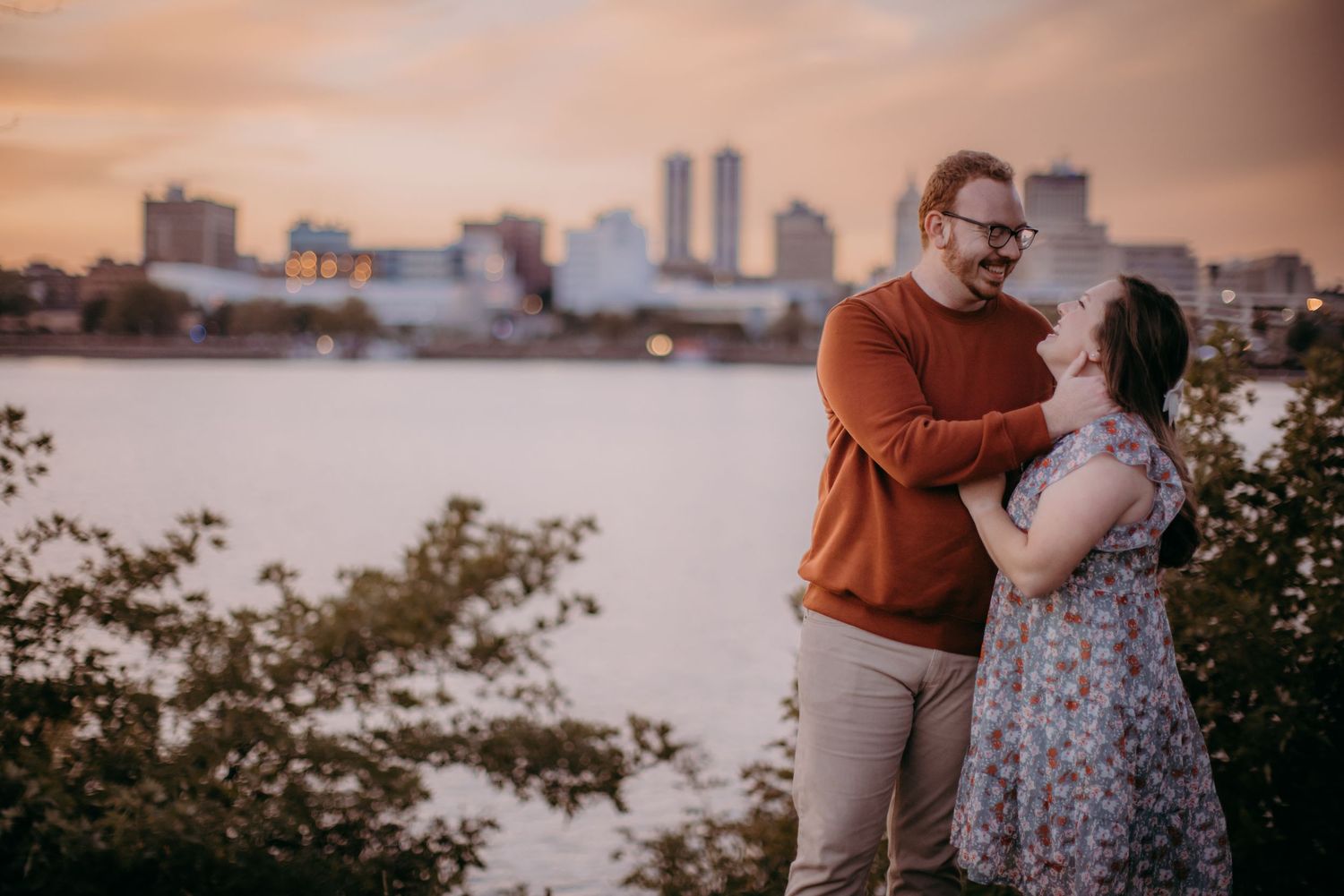 Two people share a romantic moment along the riverbank with a cityscape silhouetted in the sunset.