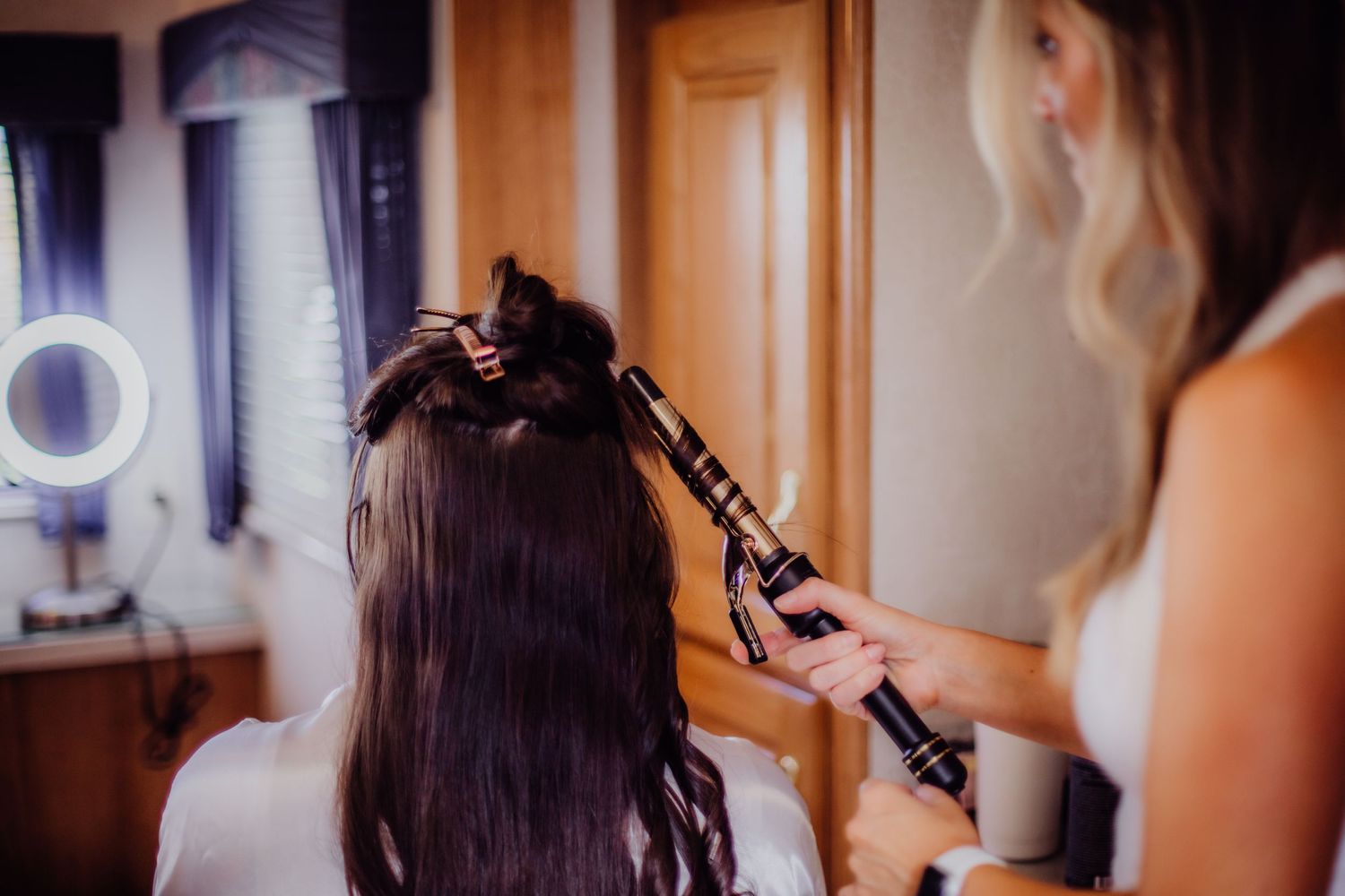 Wedding day preparation scene with hair styling and makeup in warm indoor lighting.