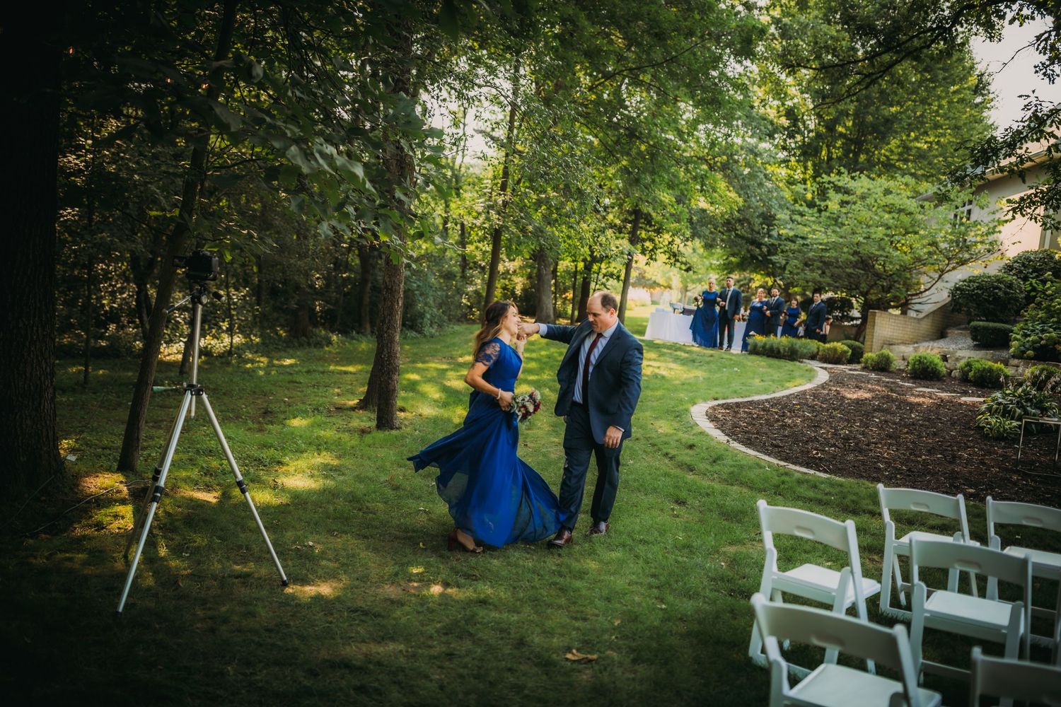 Outdoor wedding venue with white chairs and couple walking wooded path.