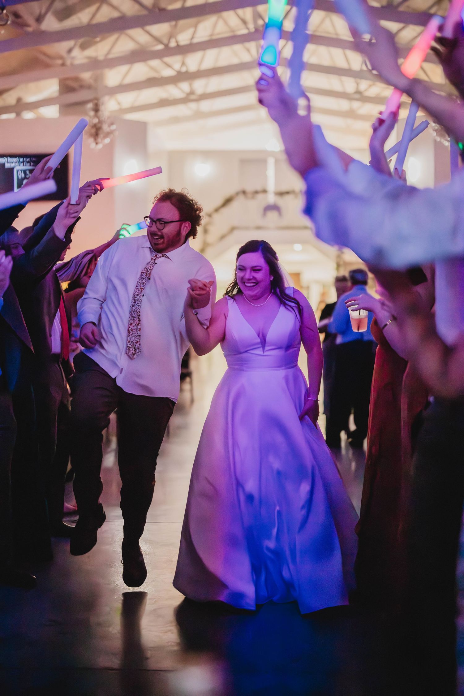Wedding couple dances through crowd of guests holding colorful light sticks at evening reception.