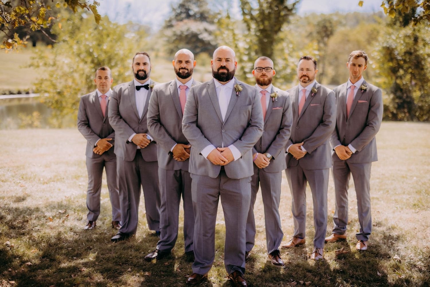 Groomsmen stand in a line wearing matching grey suits and bowties in an outdoor setting.