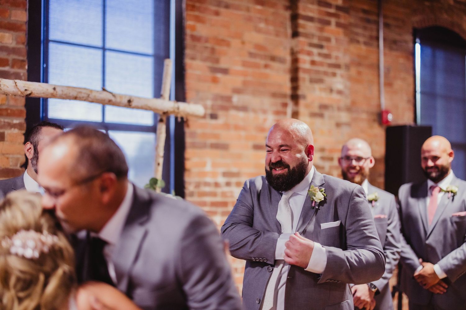 Wedding party members in gray suits share emotional moment during ceremony in industrial venue.