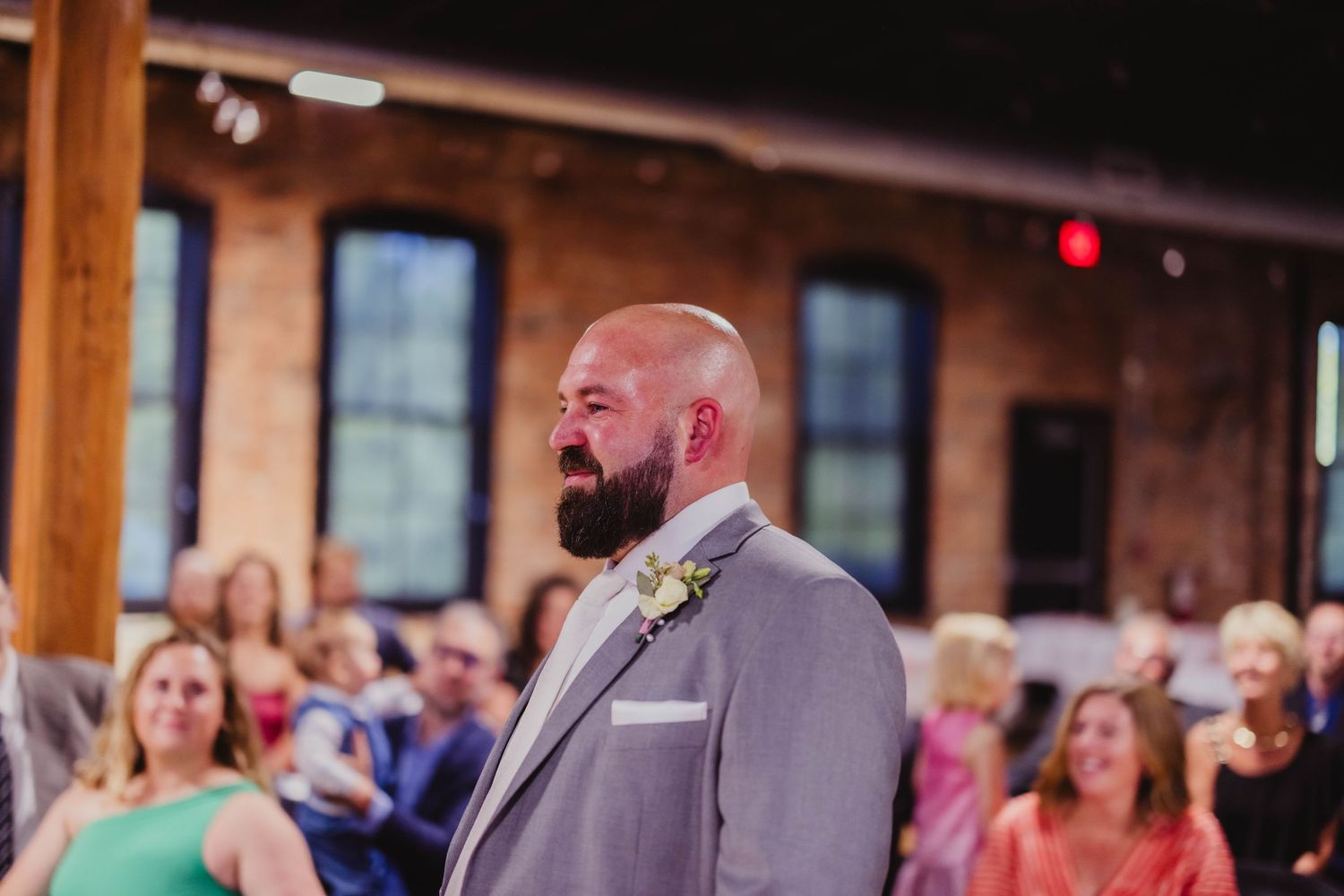 A smiling groomsman in a light grey suit stands at a wedding ceremony in an industrial-style venue.
