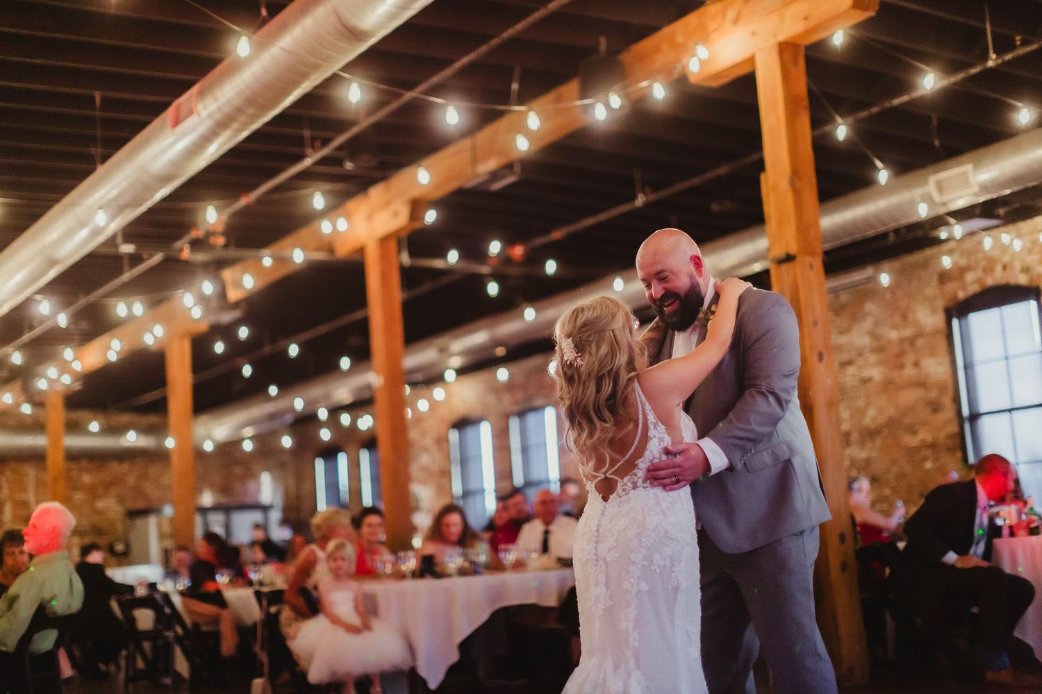 Wedding couple dances in rustic venue with exposed brick walls and string lighting.