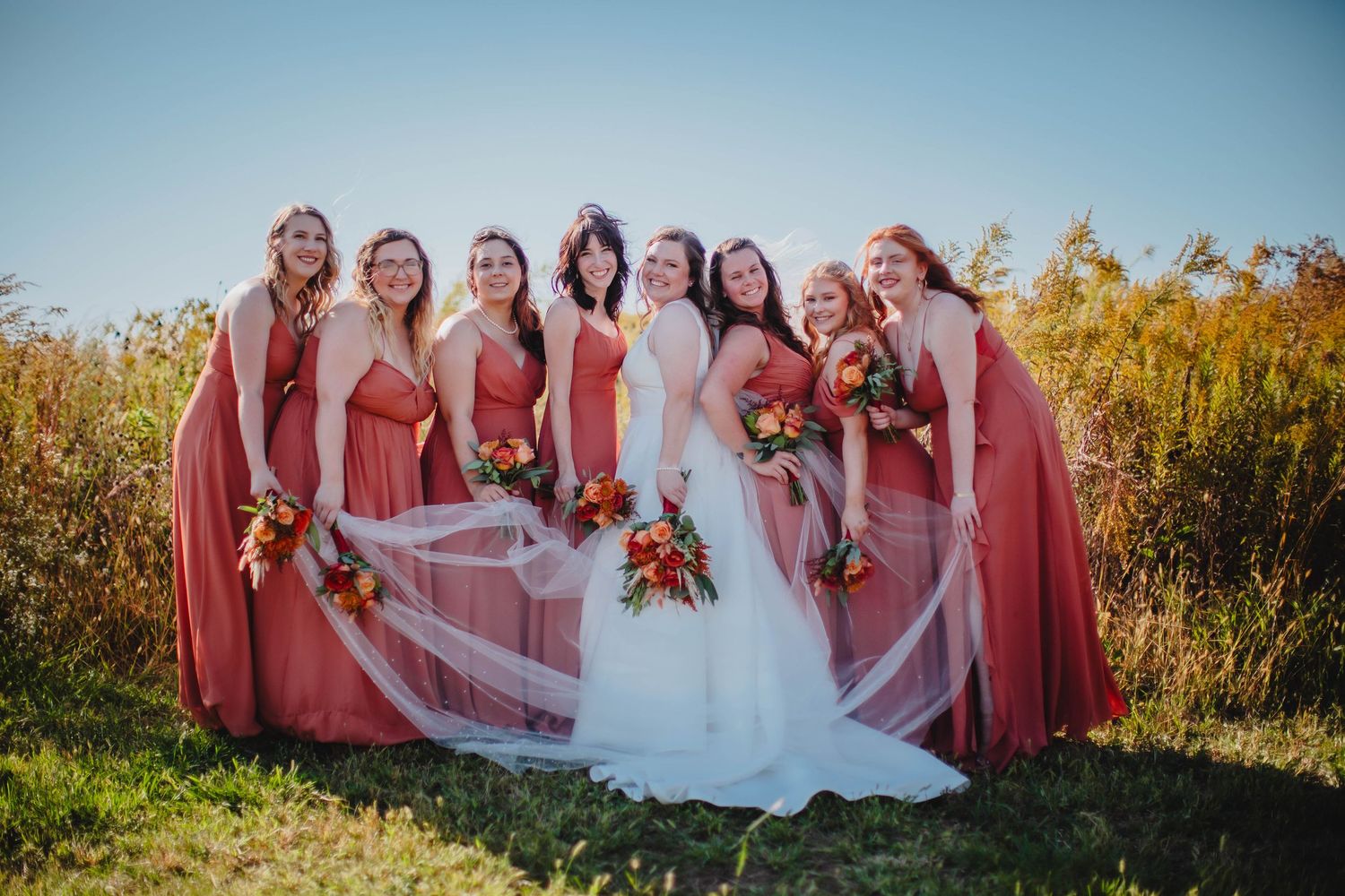 Bride's veil flows in breeze as bridesmaids in coral dresses gather for photo in autumn field.