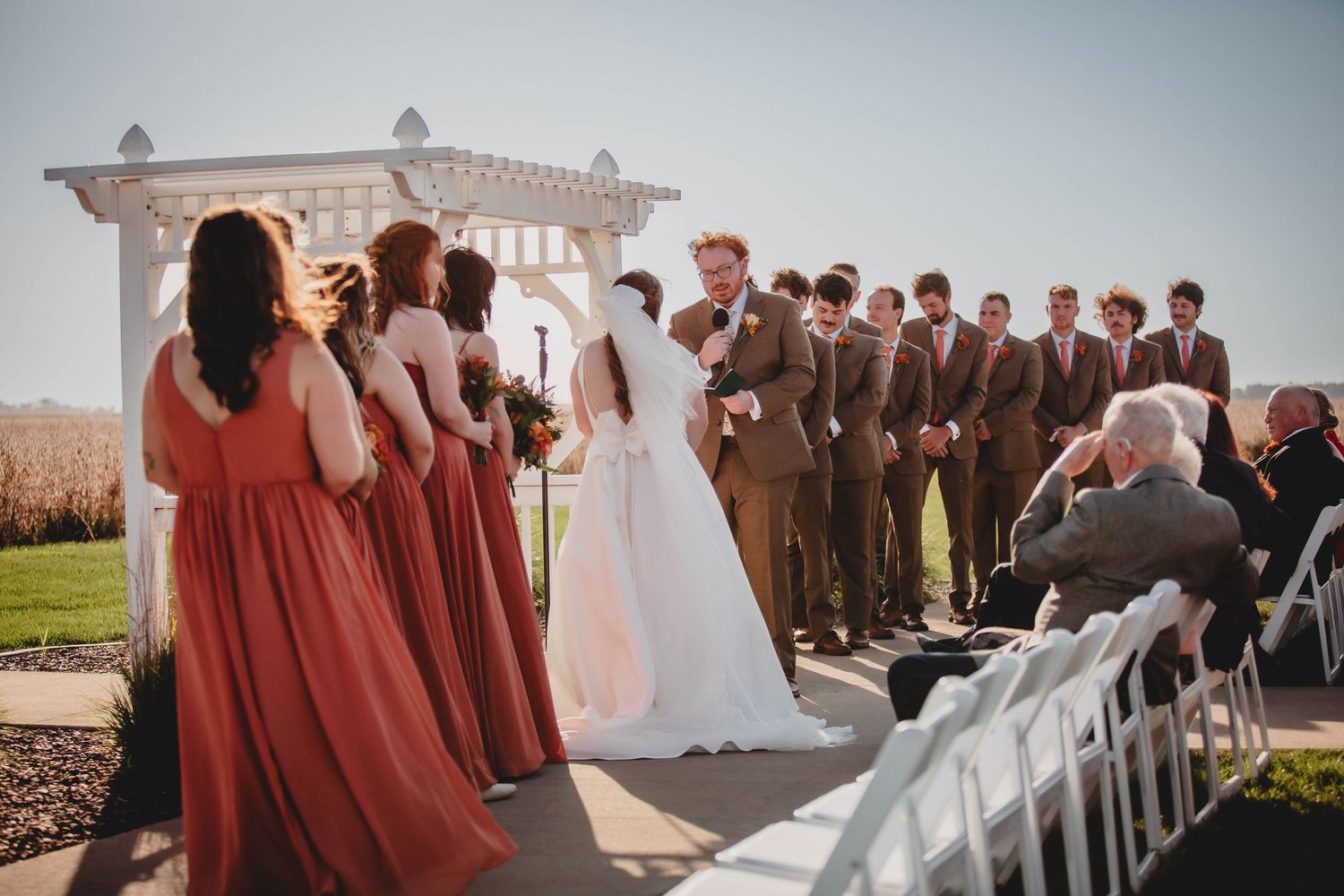 Wedding party in coral dresses and brown suits gather under outdoor pergola.
