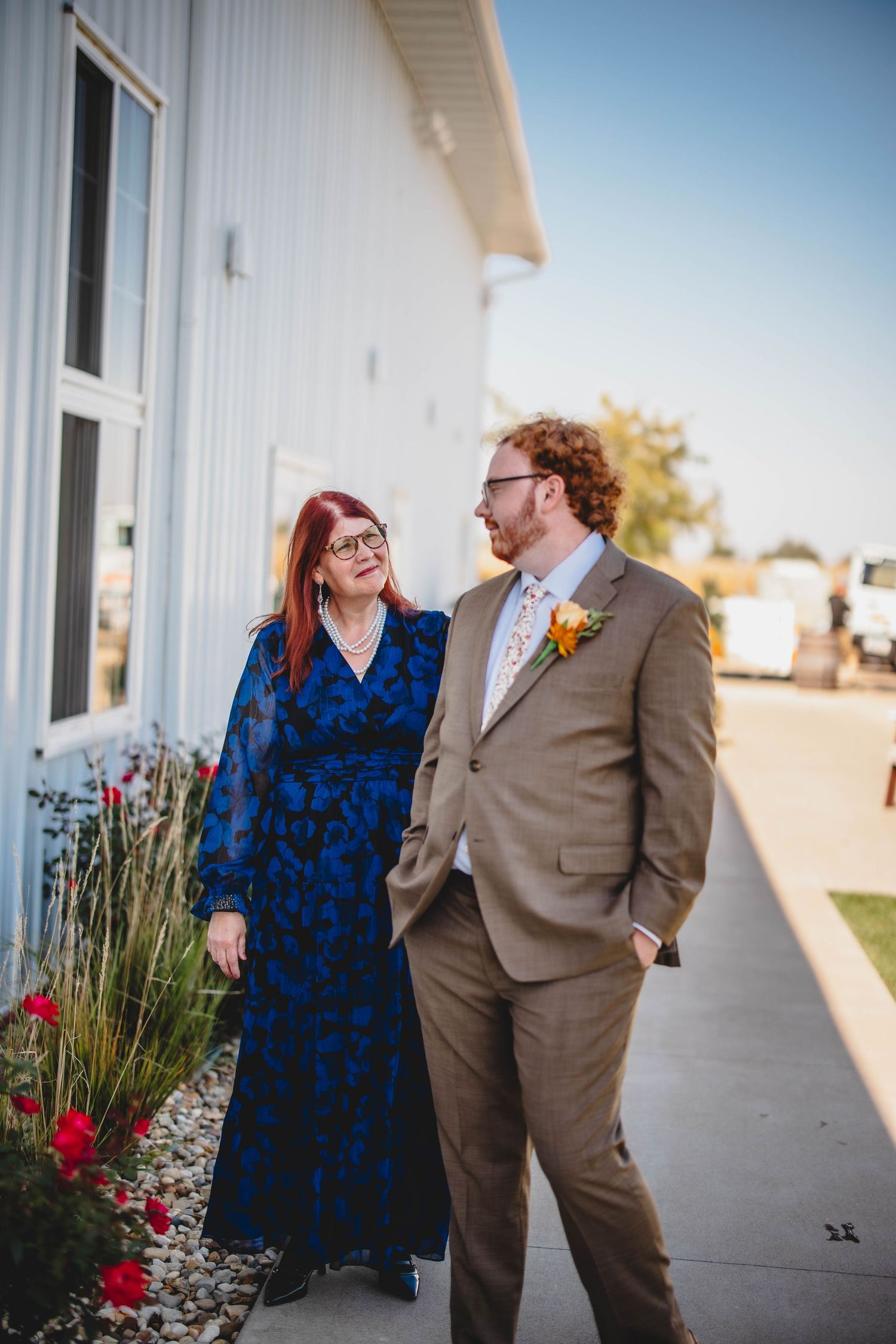 A couple in formal wear standing by a white building with red flowers.