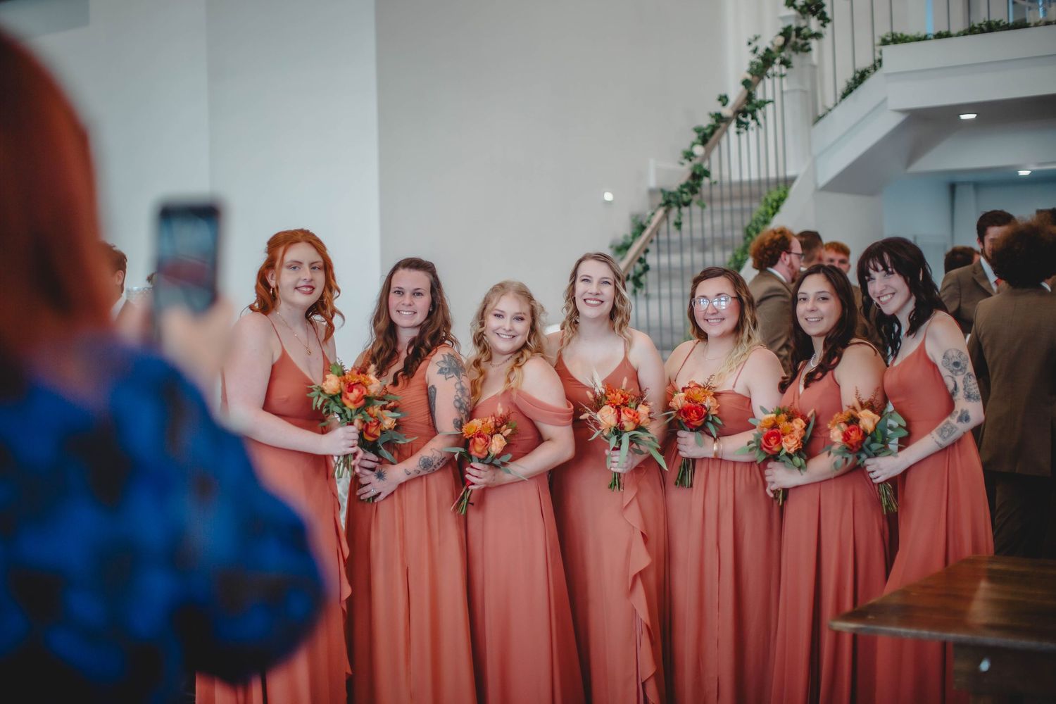 A line of bridesmaids in matching coral dresses pose together while someone photographs them.