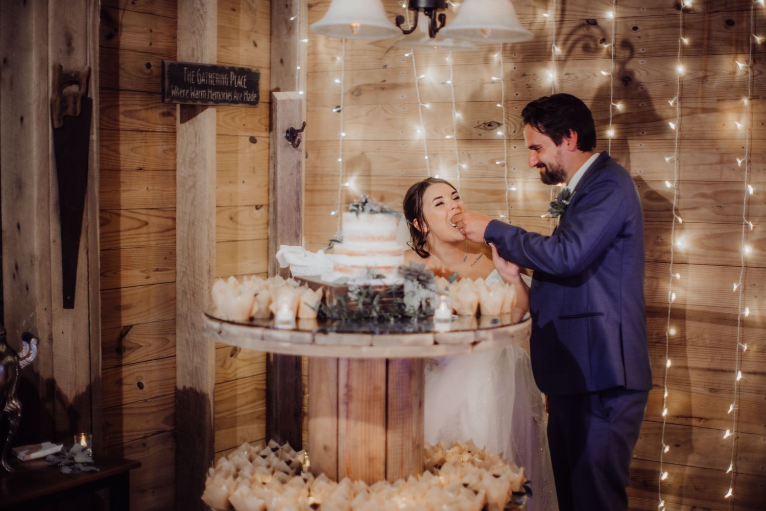 Wedding couple feeds each other cake during their reception in a barn decorated with string lights.