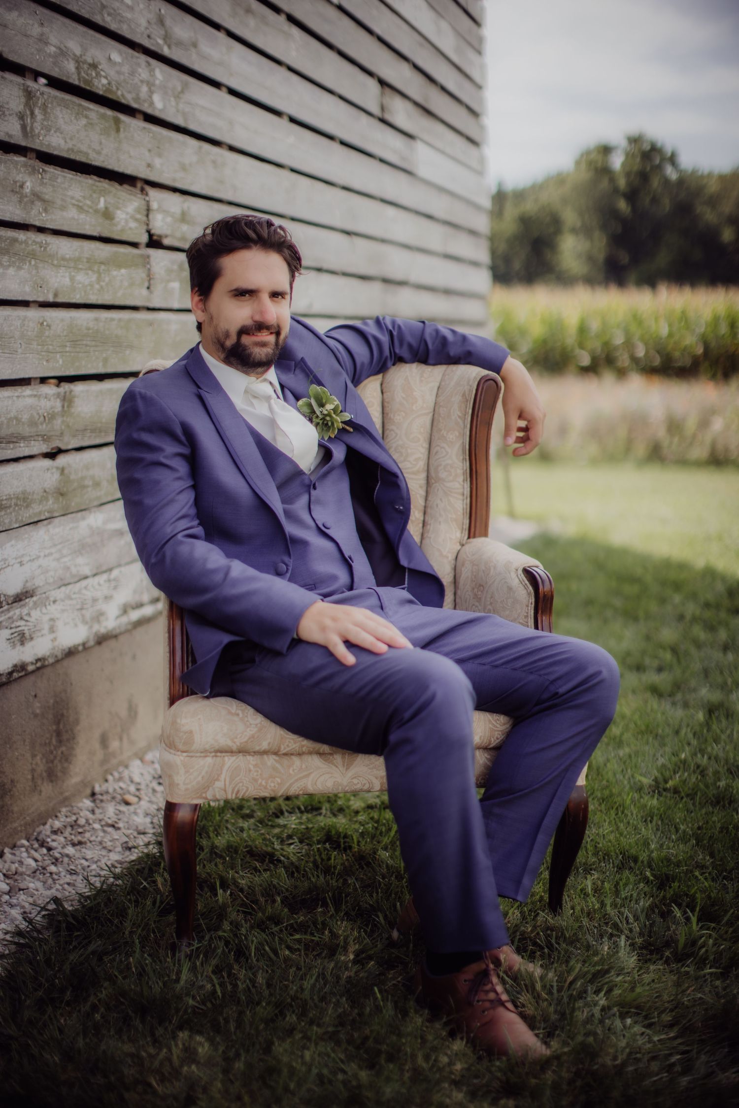 Sophisticated groom portrait in fitted navy suit relaxing in antique armchair outdoors.