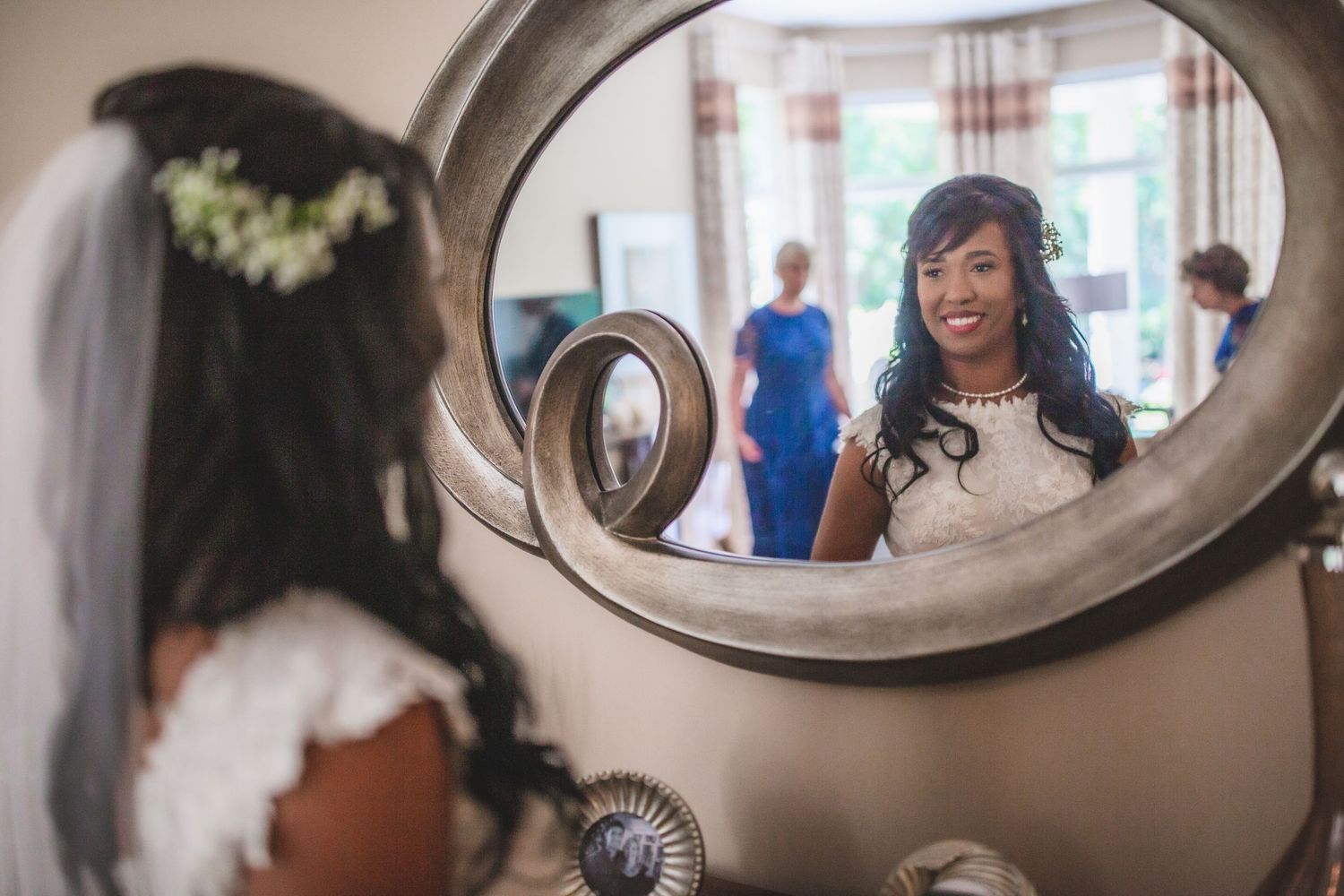 Reflection of a bride wearing a delicate veil and white dress in an ornate oval mirror.