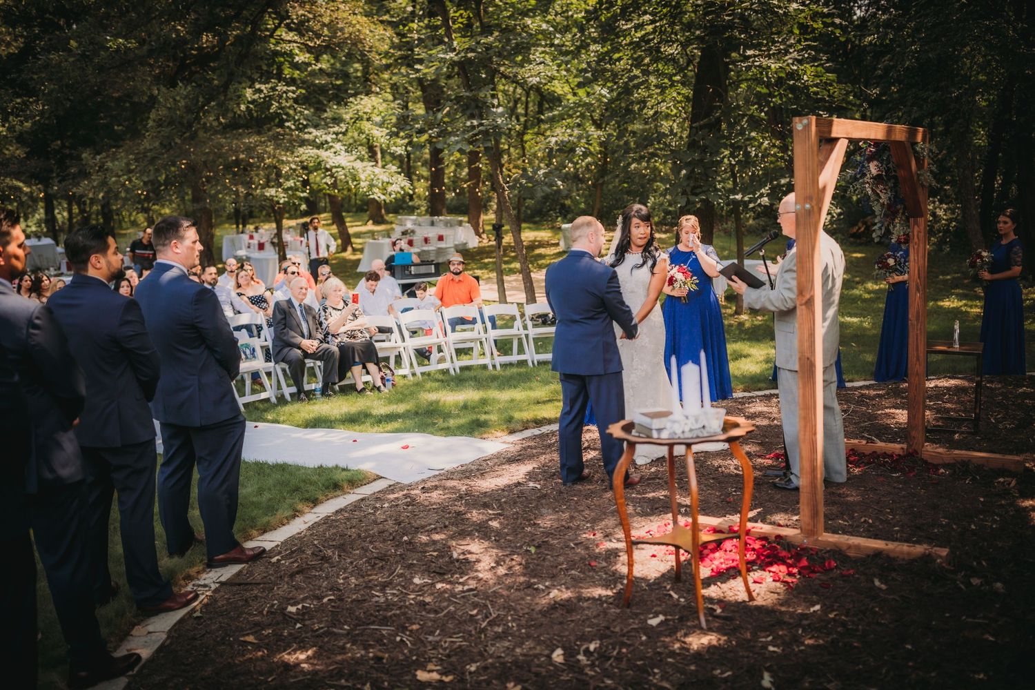 Wide angle shot of a garden wedding ceremony with guests seated beneath tall trees.