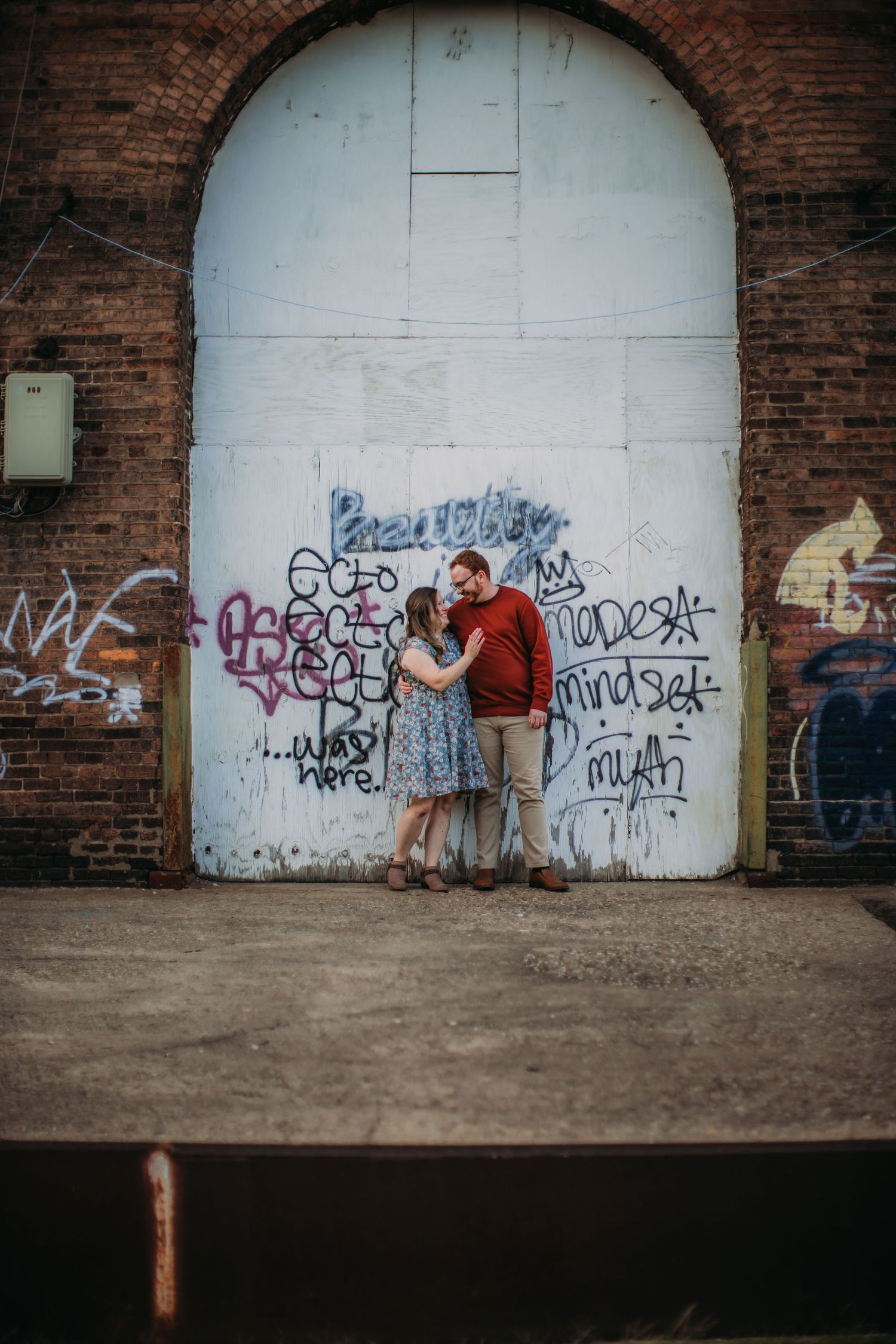 A couple poses against a graffiti-covered brick wall in an urban setting.