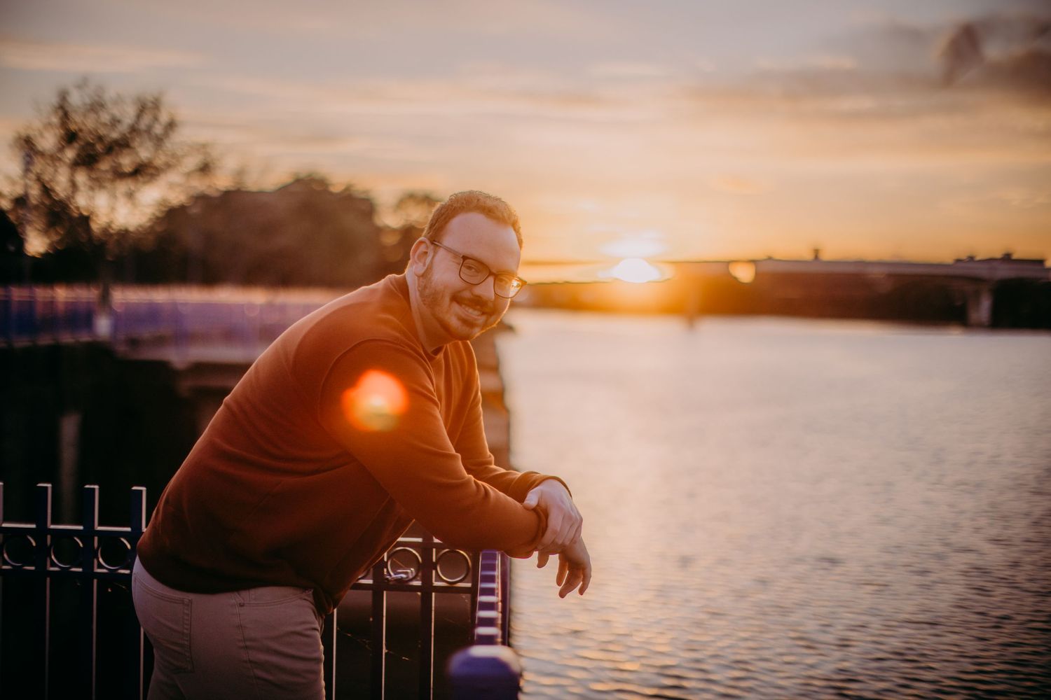 Person in casual attire leans on railing overlooking water at sunset.