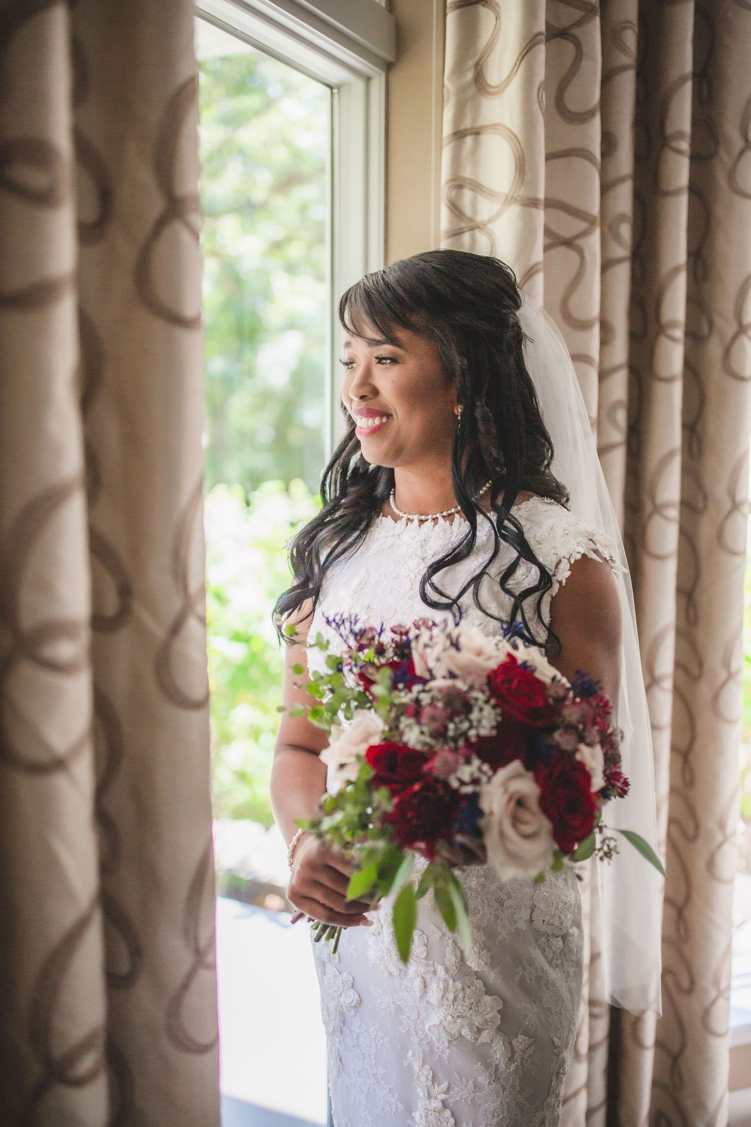 A bride in a white lace dress holds a burgundy and pink bouquet while standing by a window with patterned curtains.