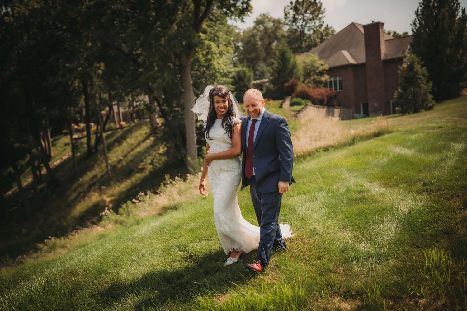 Wedding couple strolls hand in hand on a sunny hillside overlooking a brick mansion.