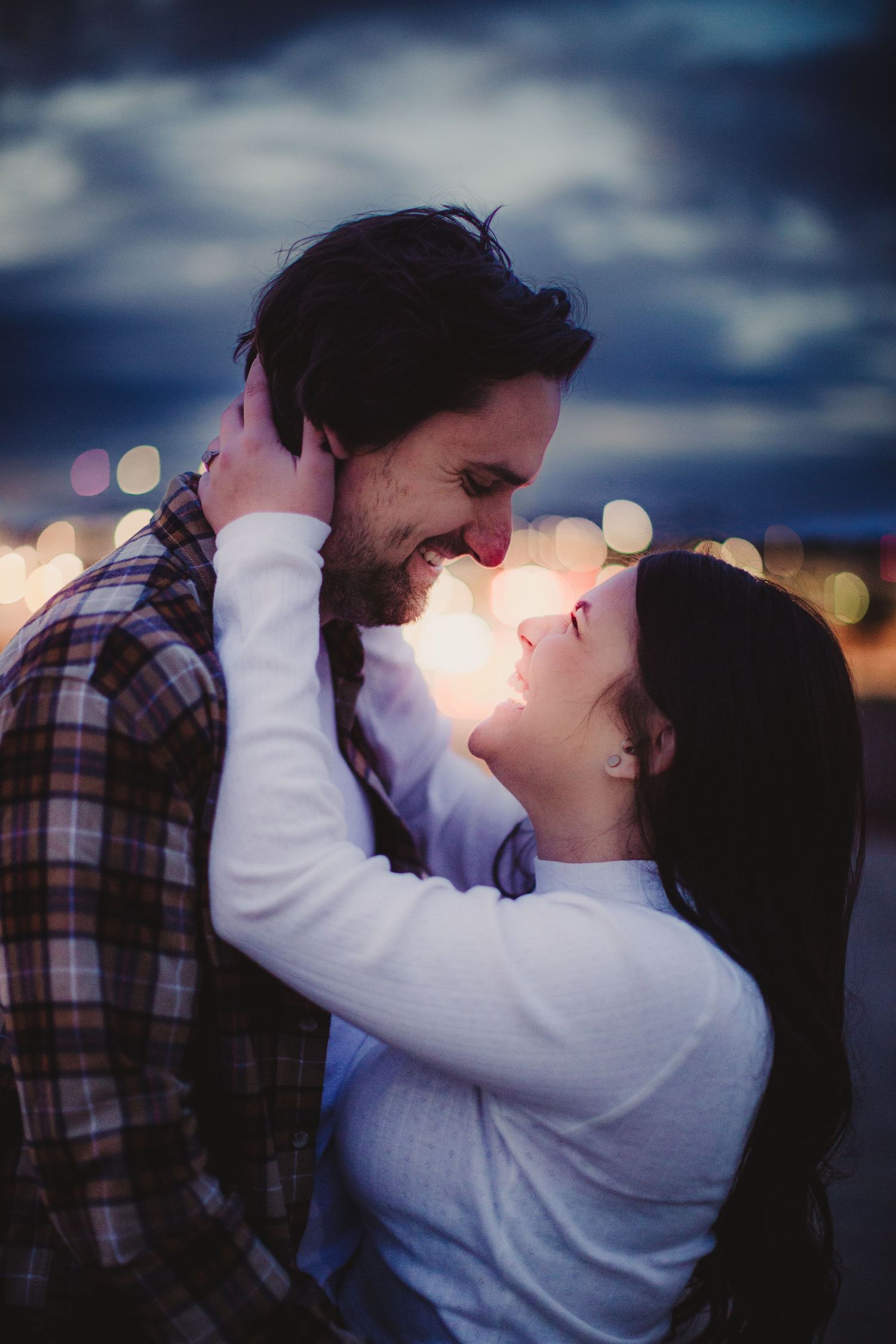 A couple shares an intimate embrace against a bokeh city backdrop at dusk.