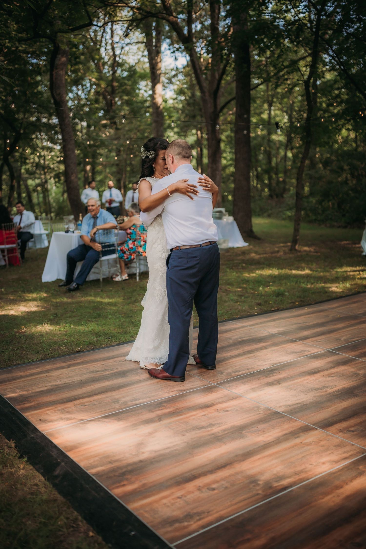 Couple embraces while dancing on a wooden platform at an outdoor wedding with guests seated among the trees.