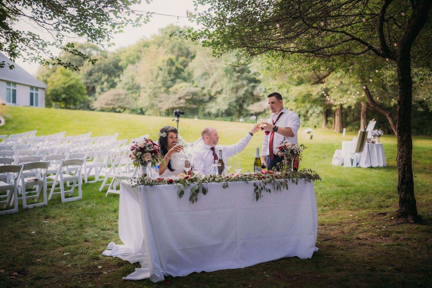 White reception tables adorned with floral centerpieces set up in a wooded outdoor venue.