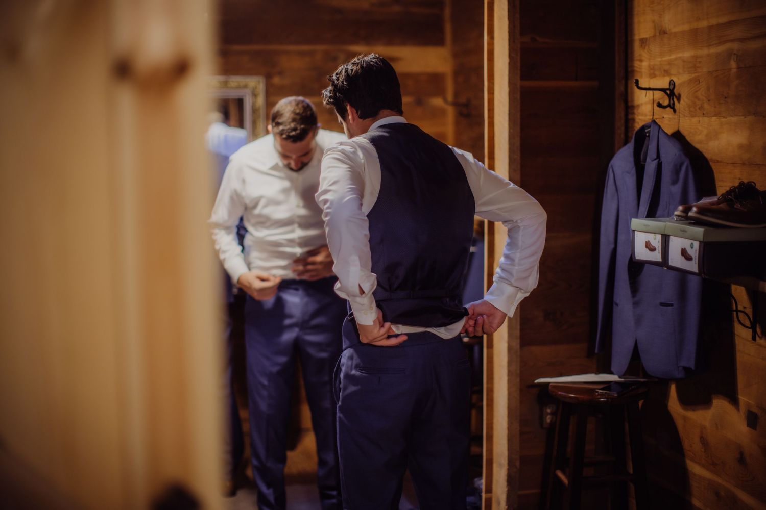 Two men in formal attire preparing in a wooden dressing room with suits hanging.