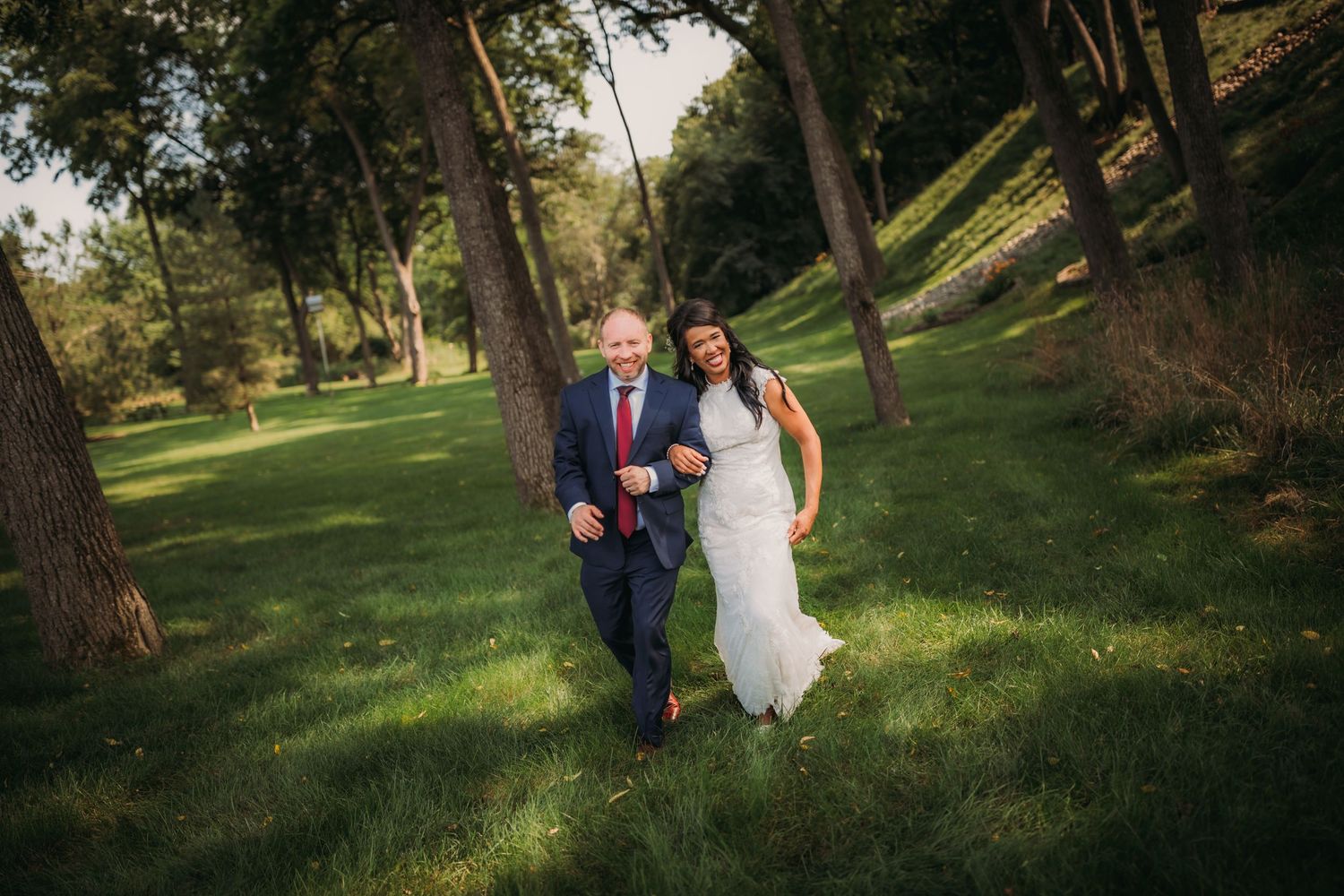A couple in wedding attire walks through a scenic park with dappled sunlight.